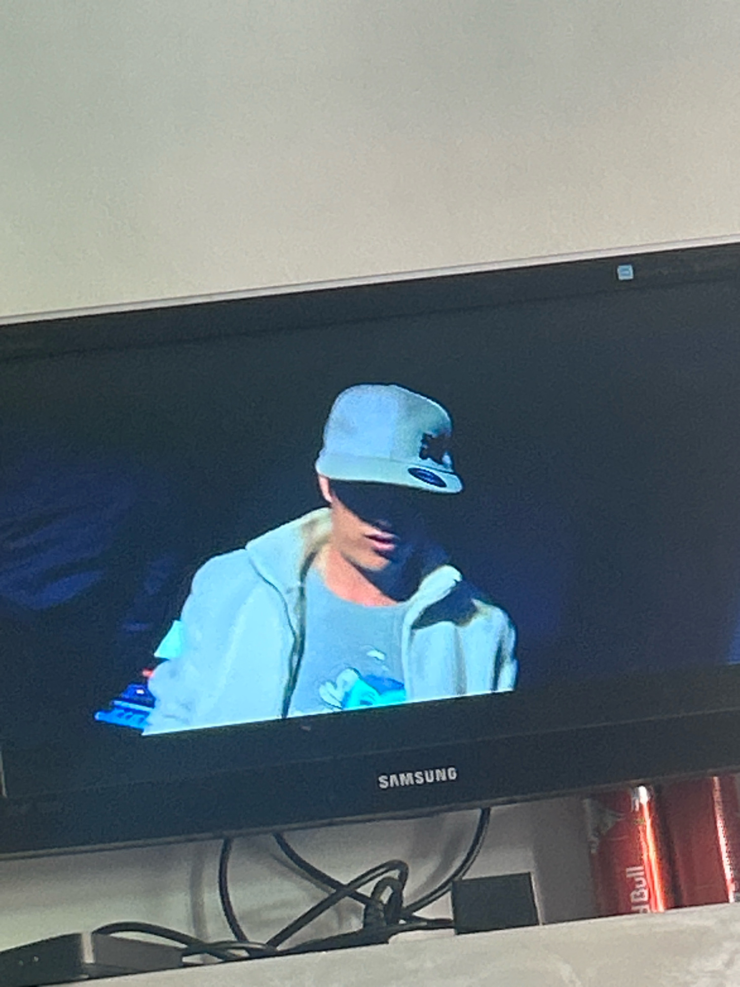 The image shows a television screen displaying a person wearing a light blue baseball cap and a light blue hooded jacket over a grey t-shirt. The person's face is largely obscured by the shadow of the cap. In the foreground, below the screen, are some electronic cables, a black rectangular object, and two cans of Red Bull. The television has "SAMSUNG" written on its frame. The image is taken from a slightly elevated angle, looking down at the television. The location is not discernible from the image itself.