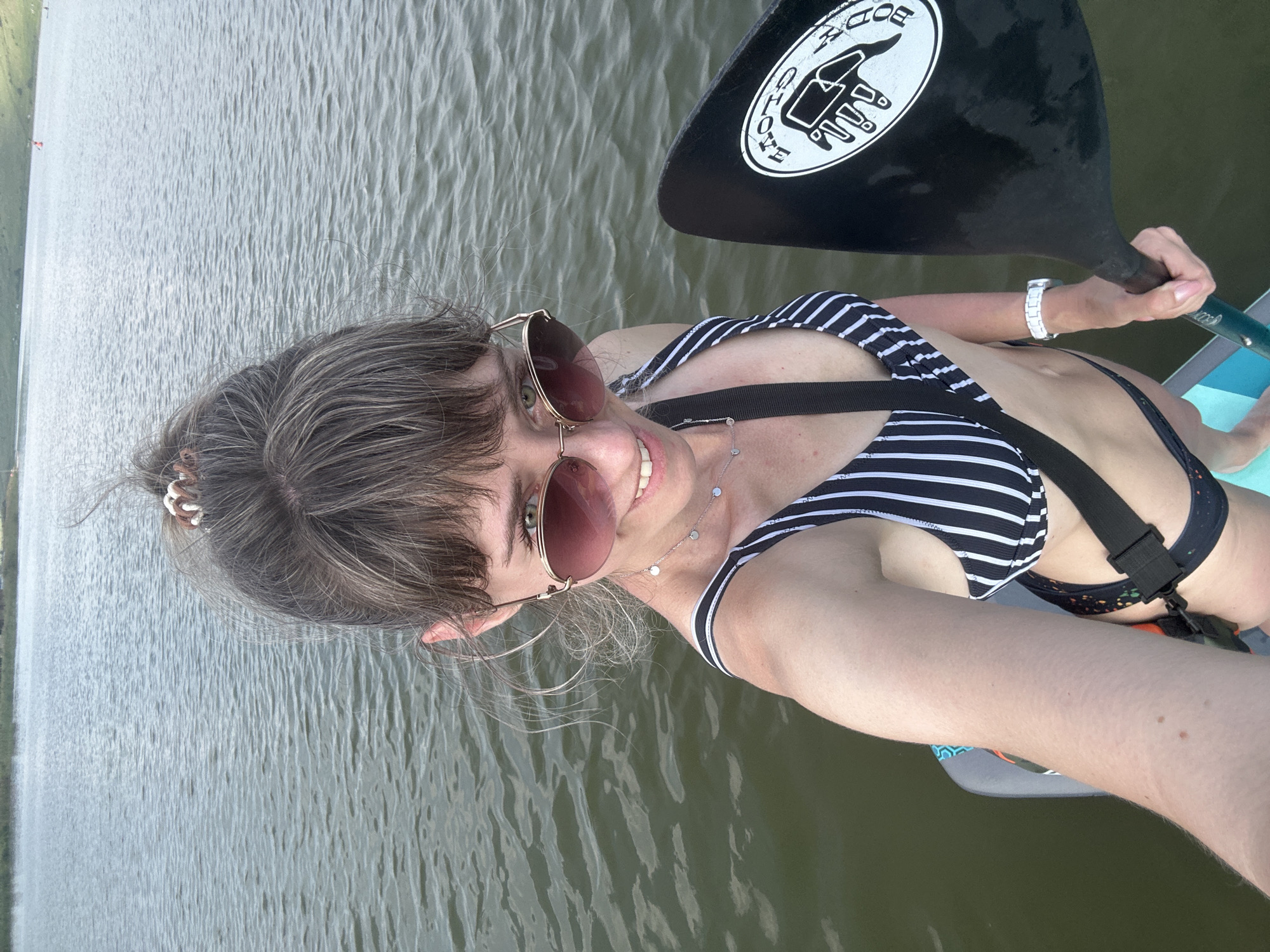 A woman is captured in a close-up, overhead selfie while paddleboarding. She is wearing a black and white striped bikini top and sunglasses with rose-tinted lenses. Her hair is pulled back with a scrunchie, and she is smiling. She holds a paddle with a logo that reads "BODY GLOVE" in white text on a black background with a white graphic. The paddleboard is visible beneath her. The background is a calm body of water, likely a lake or river, with ripples on the surface. The lighting suggests it is daytime, possibly late afternoon given the softness of the light. The woman appears relaxed and happy, enjoying the activity. There are no other people or specific landmarks visible in the frame, making the exact location indeterminate.