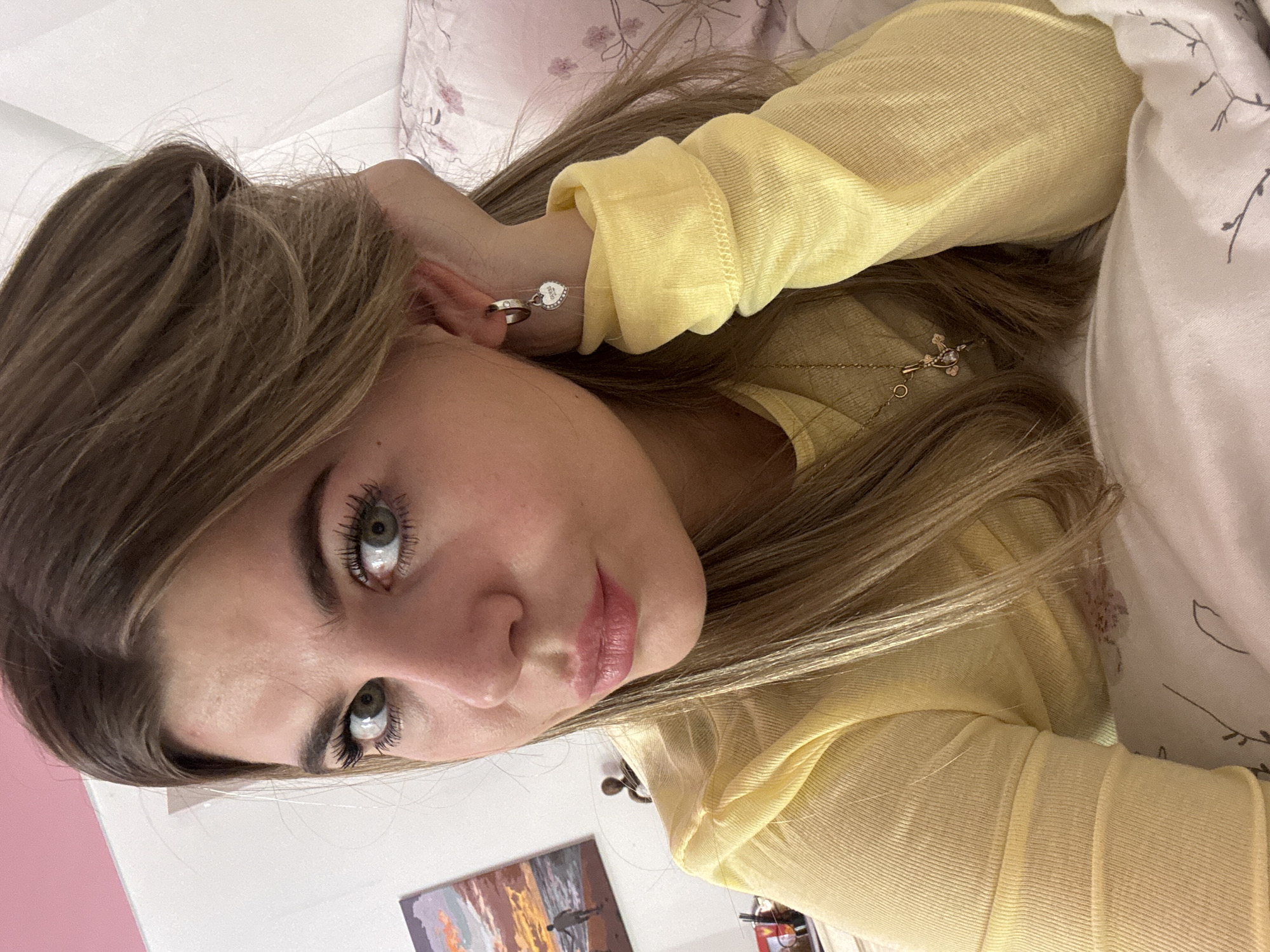 A young adult female is depicted in a close-up, selfie-style photograph, positioned indoors, likely in a bedroom. She has light skin, long, wavy light brown hair, green eyes with visible eyelashes, and pink-toned lips. She wears a ribbed, long-sleeved yellow top, a silver-toned hoop earring with a heart-shaped charm (bearing the "GUESS" brand name) in her right ear, and a delicate gold-toned necklace with a small pendant. Her right hand is positioned with the palm resting under her jawline, partially supporting her head. She looks directly at the camera with a neutral expression. The background features a plain white wall, with a small section of a pink wall visible in the upper left. A framed picture depicting a sunset or sunrise scene over water with a silhouetted figure is visible on the white wall. Below this picture, a small, dark, abstract figurine is partially visible on a surface. Floral-patterned white and pink bedding, likely a pillow or duvet, is visible on the right side of the frame. There are no discernible visual identifiers in the image to determine a specific location within the city of Chaikovsky, Russia; however, the scene is consistent with a private indoor residence.