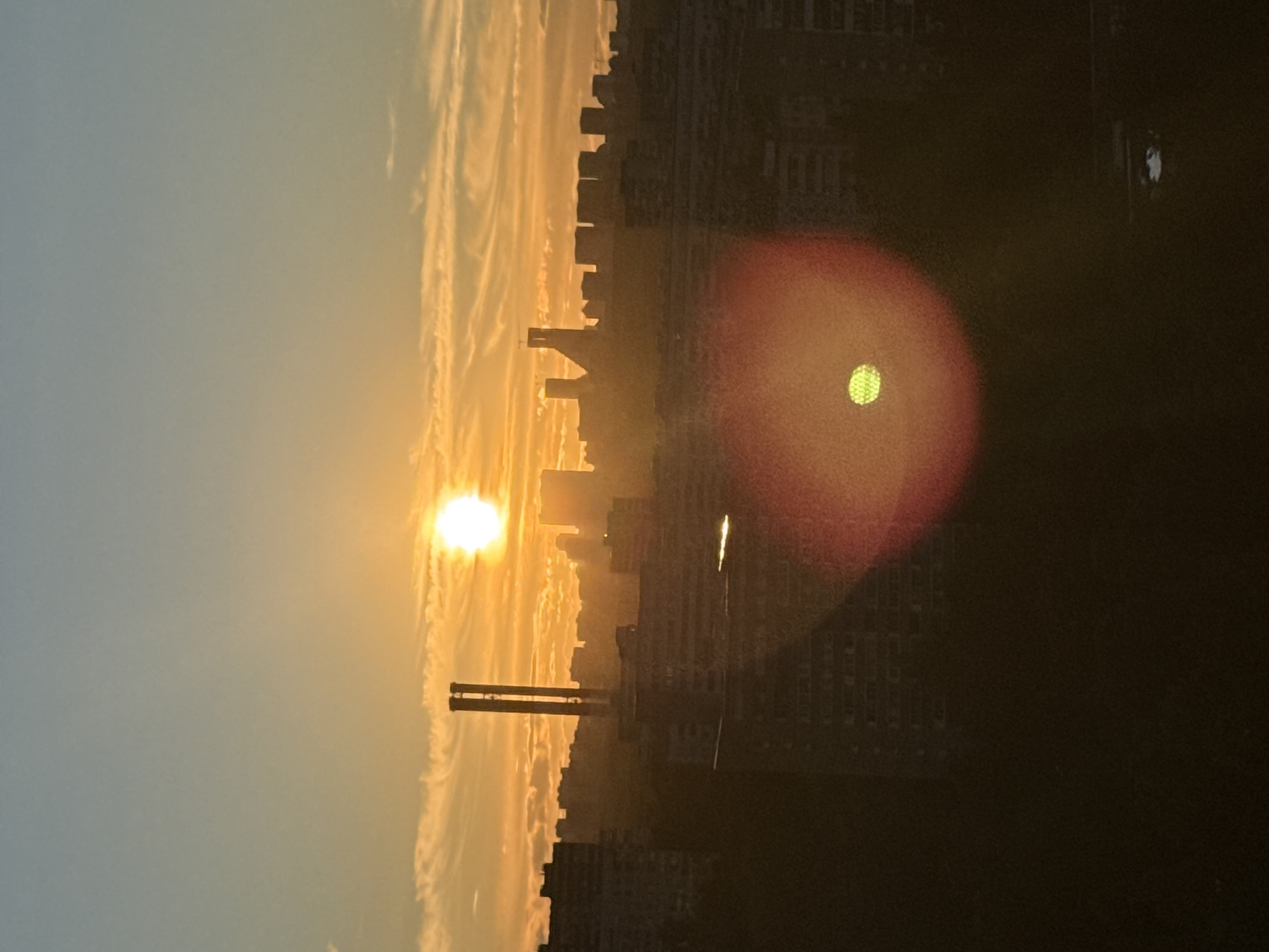The image captures a sunset over a city skyline, likely Moscow, Russia, given the context. The sun, a bright orb casting a warm golden glow, is positioned towards the left of the frame, partially obscured by wispy, elongated clouds that stretch across the sky. These clouds are illuminated by the setting sun, creating a dramatic and ethereal effect. In the foreground, silhouetted against the vibrant sky, is a cityscape featuring numerous buildings of varying heights, suggesting an urban environment. A prominent, industrial-looking structure with two tall, parallel columns stands out in the mid-ground, adding a unique element to the scene. The light from the setting sun creates a hazy, atmospheric effect, with buildings in the distance appearing softer and less defined. A reddish-orange lens flare with a green circular object within it is visible on the right side of the image, indicating the direction of the sun and the presence of a light source affecting the camera's lens. The overall mood is serene and picturesque, evoking the tranquility of a day's end. The time of day is clearly sunset due to the low angle of the sun and the characteristic warm hues of the sky. No visible text is present in the image.