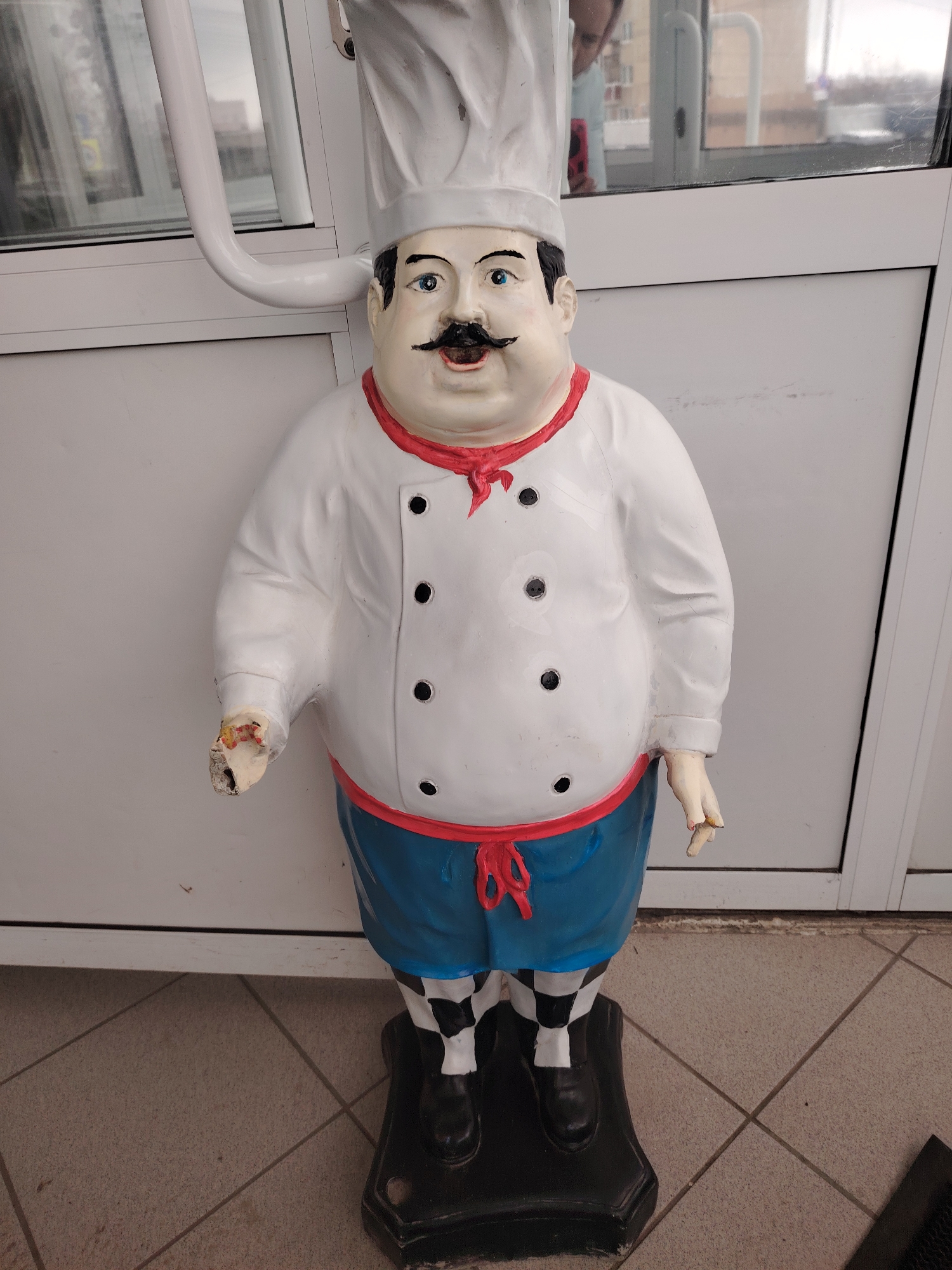 A life-sized, painted plaster statue of a male chef stands indoors on a tiled floor. The statue has a white chef's hat and jacket with black buttons, a red neckerchief, blue pants, and black and white checkered leggings. The statue rests on a black base. The statue's left hand is broken. The background consists of a white door and a window with a metal frame and handle. The location is Sarapul, Russia.