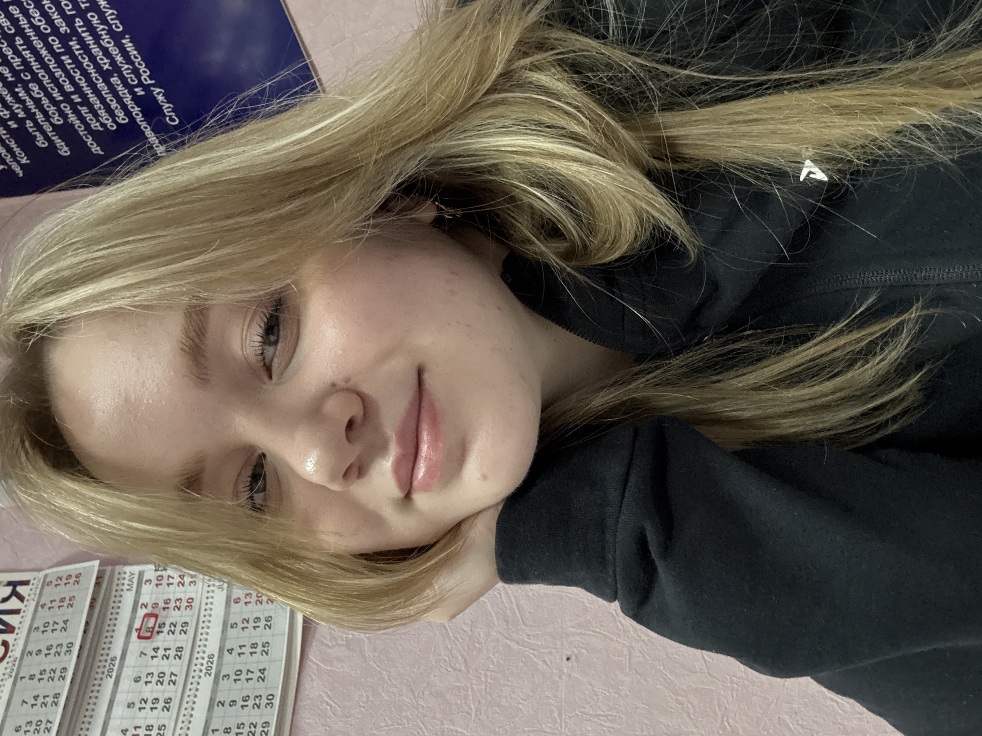 The image depicts a young woman with blonde hair resting her head on her hand, likely on a pink surface. She is wearing a black hoodie. A calendar is visible, showing the months of April and May 2026. The printed text above the calendar indicates a location in Russia, mentioning the phrase "Служу России" (I serve Russia). Based on the other text in the image, the location may be Chaikovsky, Russia.