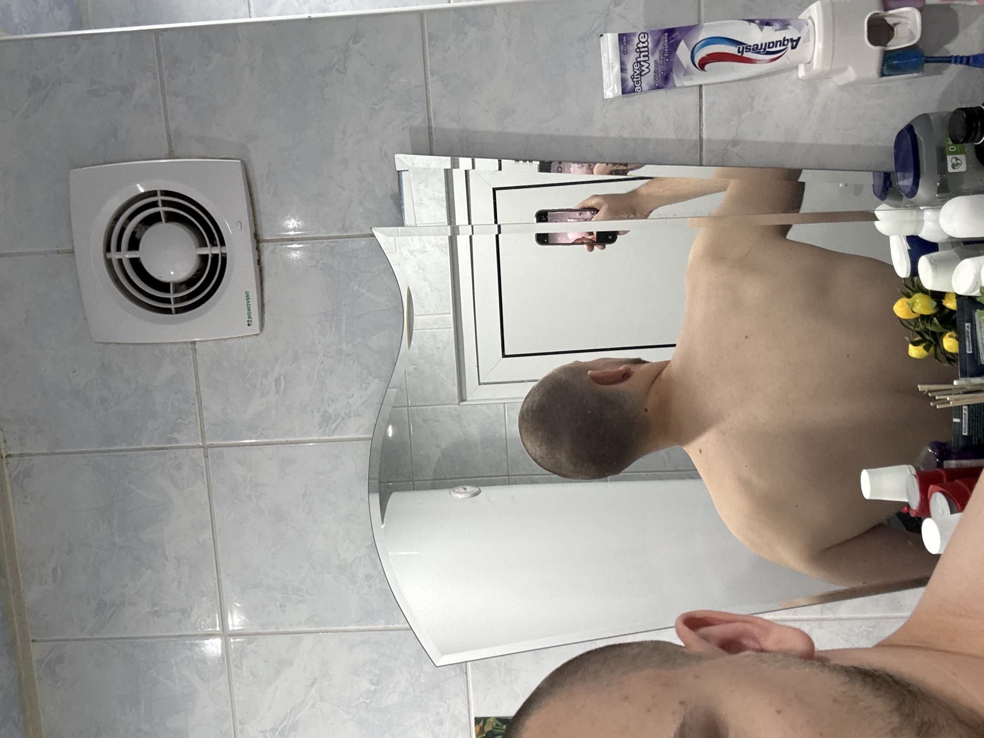 The image displays a bathroom interior in Stara Zagora, Bulgaria, captured as a mirror selfie. The room's walls are covered with light grey, slightly mottled square tiles with matching grout.
In the center of the frame is a multi-panel mirror with decorative borders, showing the reflection of a person taking the photograph. The person, appearing to be male with a very short haircut and visible stubble/beard, is seen from the top of the head (forehead, eye area, ear) in the foreground, and his bare back and shoulder are reflected further into the mirror. He is holding a black smartphone with a bright screen to capture the image. The mirror's segmented design creates vertical lines and slight distortions in the reflection.
To the left, mounted on the tiled wall, is a white square ventilation fan with a circular grille, bearing the green text "DOMOVENT". Above the mirror, a white toothpaste dispenser holds a tube of "Aquafresh active White" toothpaste. Visible text on the toothpaste includes "WALSON", "whitens", "cleans", and "freshens". To the right, on a shelf or counter next to the mirror, are various bathroom products such as bottles with white pumps or caps, and a small decorative bunch of artificial yellow flowers. The lighting in the bathroom appears bright and even.