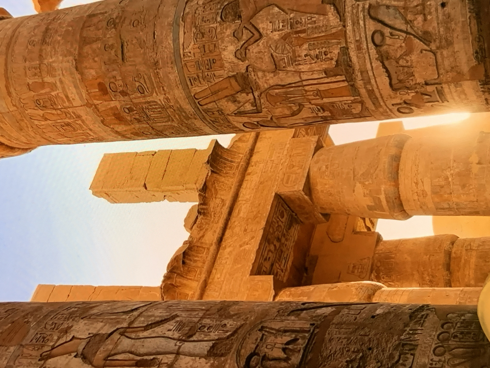The image shows a close-up view of massive, ancient Egyptian-style stone columns with hieroglyphic carvings. The columns are illuminated by warm, golden sunlight, suggesting it is either sunrise or sunset. The sky in the background is a pale blue, transitioning to a lighter hue near the horizon. Architectural elements, including lintels and other stone structures adorned with similar carvings, are visible between and behind the columns. The style and carvings are distinctly ancient Egyptian. The image is not located in Stavropol, Russia; it depicts an ancient Egyptian site.