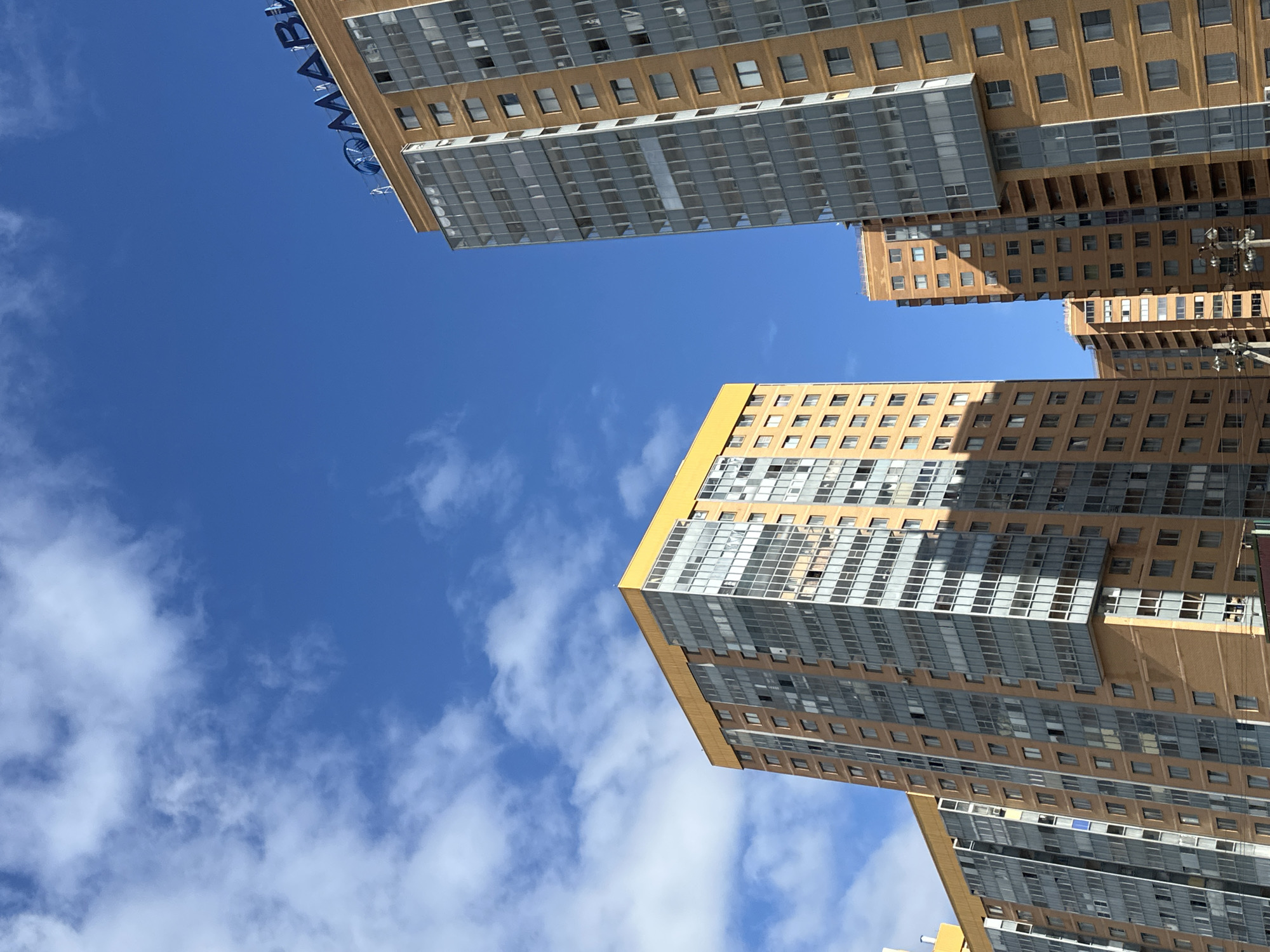 In Murino, Russia, under a bright blue sky with scattered white clouds, several modern multi-story residential buildings are visible. The buildings are constructed with light brown brick facades and feature extensive glass balconies and windows. One of the taller buildings on the right has blue neon lettering at its apex that reads "МАВИ". The perspective is from a low angle looking upwards at the architecture. The environment appears to be a residential urban setting.