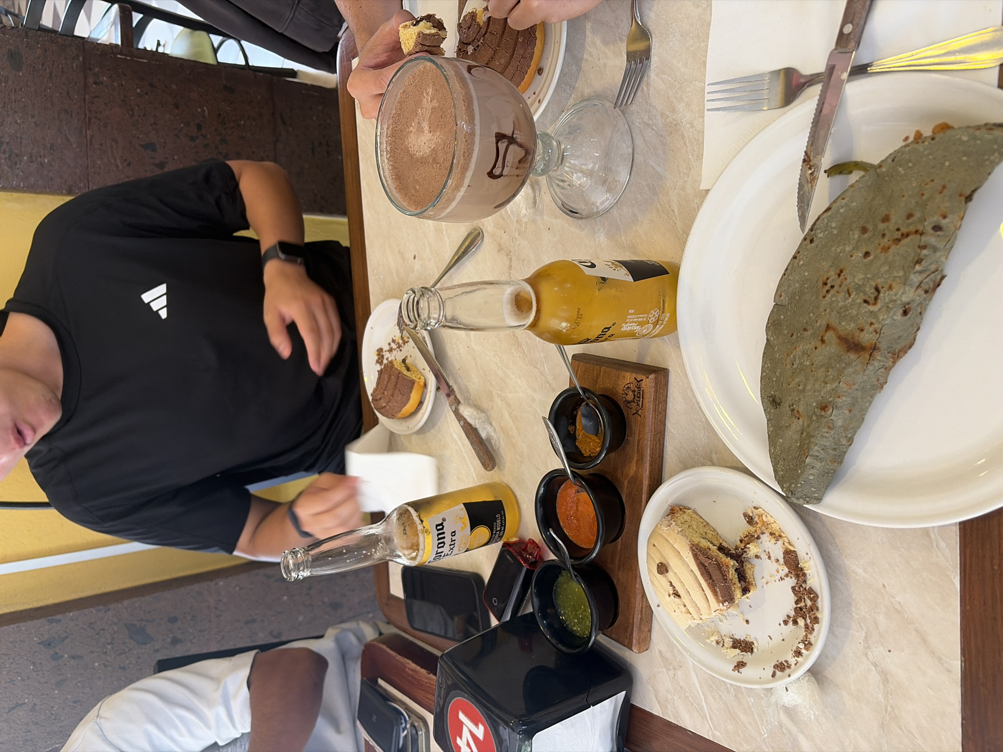 The image shows a table with various food and drink items, and two individuals. One person, wearing a black Adidas shirt, is partially visible on the left side of the image. Another person is visible at the top, holding a piece of cake, and eating with a fork. The table holds two plates of food, one containing what appears to be a blue corn quesadilla, and the other containing a concha pastry that has been partially eaten. There is also a dish of cake, a glass of chocolate beverage, and a wooden tray holding three small bowls containing different types of salsa. Two Corona beer bottles are visible, one empty, and a cell phone and an electronic cigarette. The location is unknown.