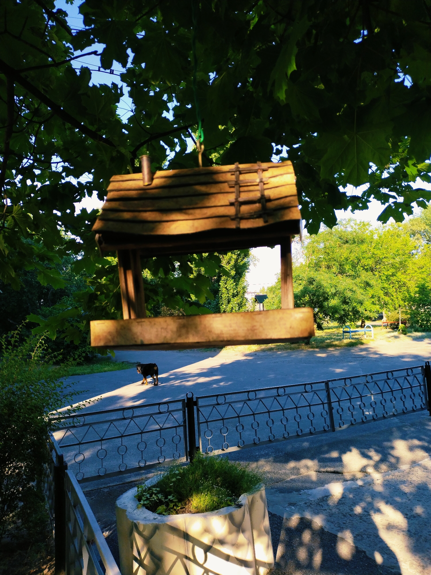A wooden bird feeder hangs from a tree branch in the foreground of this image, casting shadows on a paved area below. A small, dark-colored dog stands on the paved area, facing away from the camera and towards the right. The bird feeder is simple in design, with a sloped roof and open sides. Behind the bird feeder, lush green trees and foliage create a dense background. A black metal fence runs horizontally across the middle ground, separating the paved area from a walkway or park area. In the distance, there are more trees and a hint of buildings. The scene appears to be in an outdoor public space, possibly a park or residential area, under bright sunlight. The location is identified as Horishni Plavni, Ukraine.