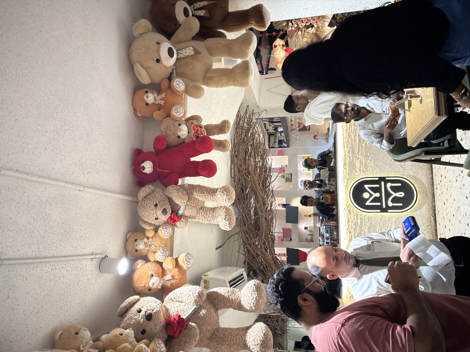 The image depicts the interior of a commercial establishment, likely a cafe or restaurant, located in Kalyan-Dombivli, India. The scene features multiple individuals and a distinctive decor dominated by stuffed teddy bears.
In the foreground, two men are engaged in a transaction. On the left, a man with a beard and glasses, wearing a salmon-colored t-shirt, holds a smartphone or similar device in his hands. Facing him, a man with a bald head, wearing a white collared shirt and a dark tie, holds a white paper receipt and a blue payment terminal. Behind them, a prominent illuminated logo, consisting of stylized interlocking 'M' shapes, is mounted on a light-colored, textured wall.
To the right, a man with glasses and a short beard, dressed in a light-colored checkered shirt, is seated at a light-colored wooden table with a beverage glass, looking towards the left. Partially obscuring him, a person with long dark hair, seen from behind, wears a black top and blue jeans.
In the mid-ground, a brightly lit bar counter is visible, behind which four individuals are present. Three staff members in dark uniforms are engaged in preparing drinks, with bottles, glasses, and a coffee machine visible on the counter. Another individual in a white hoodie stands behind the counter, looking right.
The upper portion of the scene is heavily decorated with numerous stuffed teddy bears of various sizes and colors (light brown, dark brown, red, orange), suspended from the ceiling or placed on elevated ledges. Several bears hold small red objects, such as roses or hearts labeled "LOVE." Below these decorations, a lattice-like structure made of intertwined branches or twigs spans across the ceiling. A white wall-mounted air conditioning unit is visible on the upper left, beneath some teddy bears. The floor is a light-colored terrazzo.