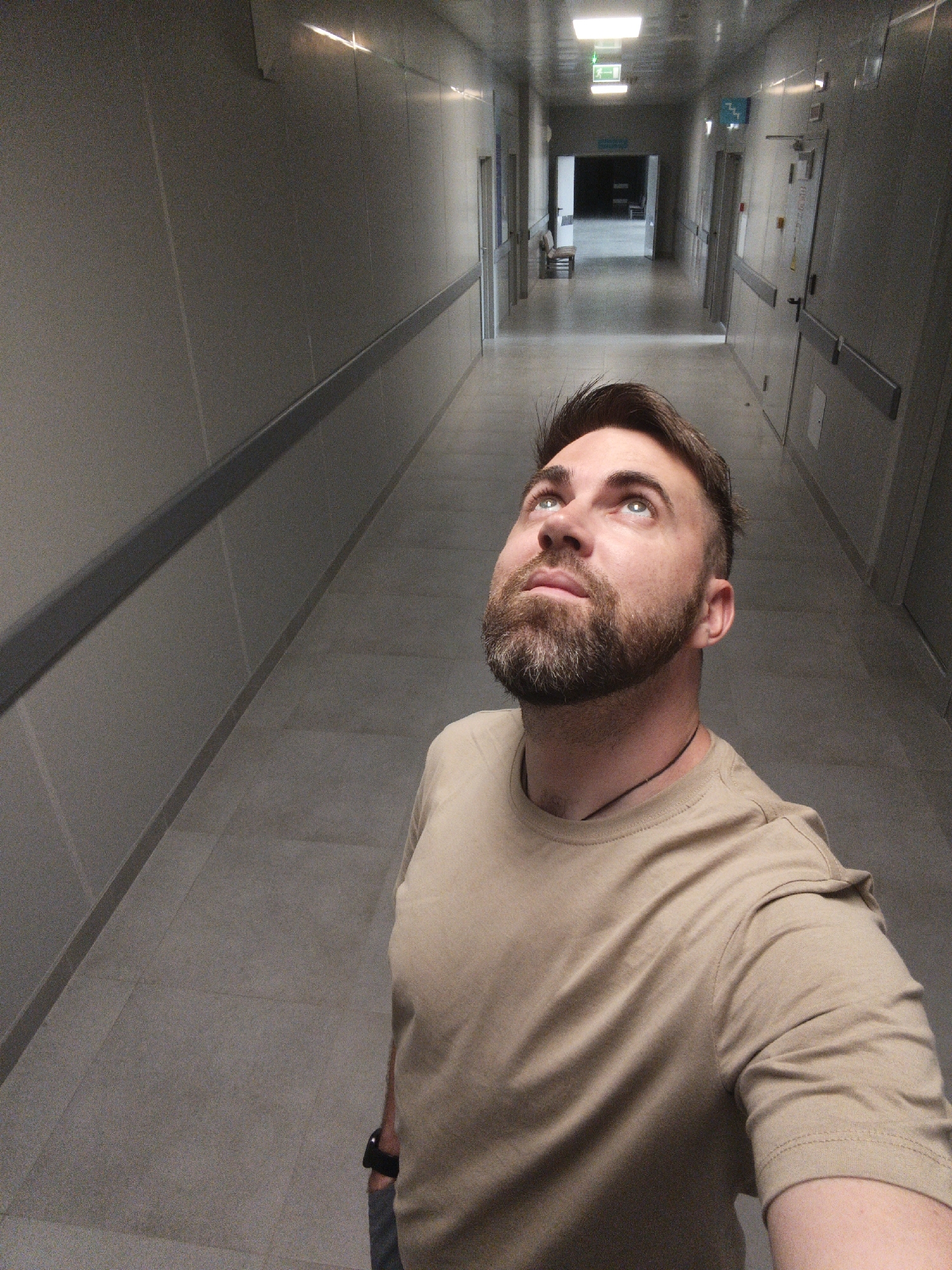 A man with a beard is standing in a long hallway, looking up. He is wearing a tan t-shirt and a thin necklace. The hallway has gray walls, gray floors, and a gray handrail. There are doors on the right side of the hallway. There are ceiling lights in the hallway. There is a dark area at the end of the hallway. The location is Балаклавский округ, Ukraine.