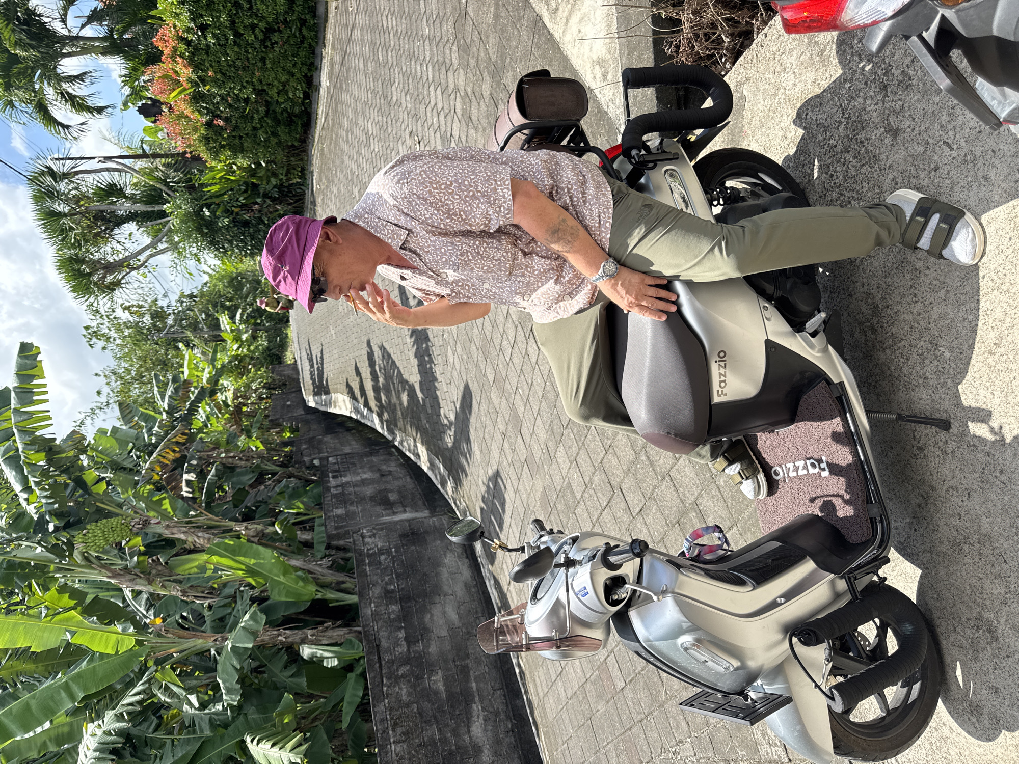 A man in a pink bucket hat, patterned short-sleeved shirt, and olive-green pants is straddling a silver scooter with "Fazzio" printed on the floor mat. He is looking down and holding something up to his face. The scooter is parked on a paved surface next to a concrete wall with lush green vegetation behind it. In the background, there are more trees and a glimpse of a building. The image appears to be taken outdoors in daylight. The location is Kediri, Indonesia.