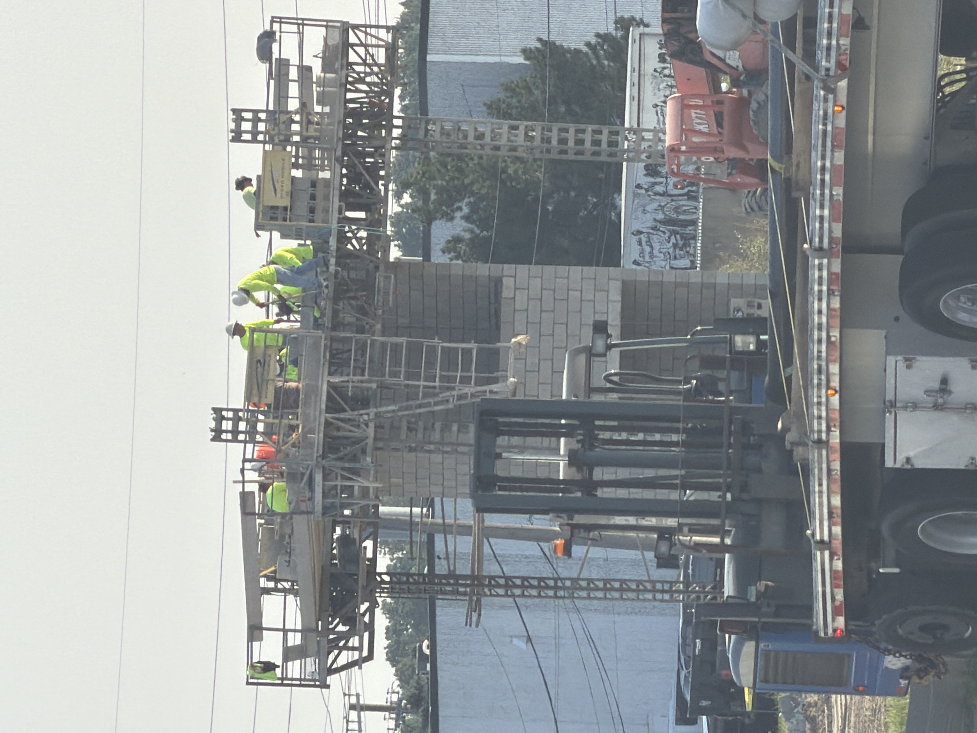 In Shelby Charter Township, Michigan, United States of America, a construction scene unfolds with workers on a platform above a brick structure. Five individuals, all wearing high-visibility safety vests, are actively engaged in their tasks on the elevated platform. Two workers are dressed in yellow vests, while three are in green vests. One worker in a yellow vest is leaning over, appearing to be working with tools, while another in a green vest is also bent over, potentially operating machinery or handling materials. The platform itself is a complex structure of scaffolding and beams, with a ladder providing access. Below the platform, a large forklift is positioned prominently, its mast raised, indicating it might be in use or ready for operation. In the foreground, a flatbed truck carries several long, dark pipes secured for transport. Behind the forklift and truck, a red piece of heavy machinery, possibly a boom lift, is visible, and further back, a white trailer with an image of people is parked. The overall environment suggests an active construction site, with buildings and trees forming the backdrop under a clear sky.