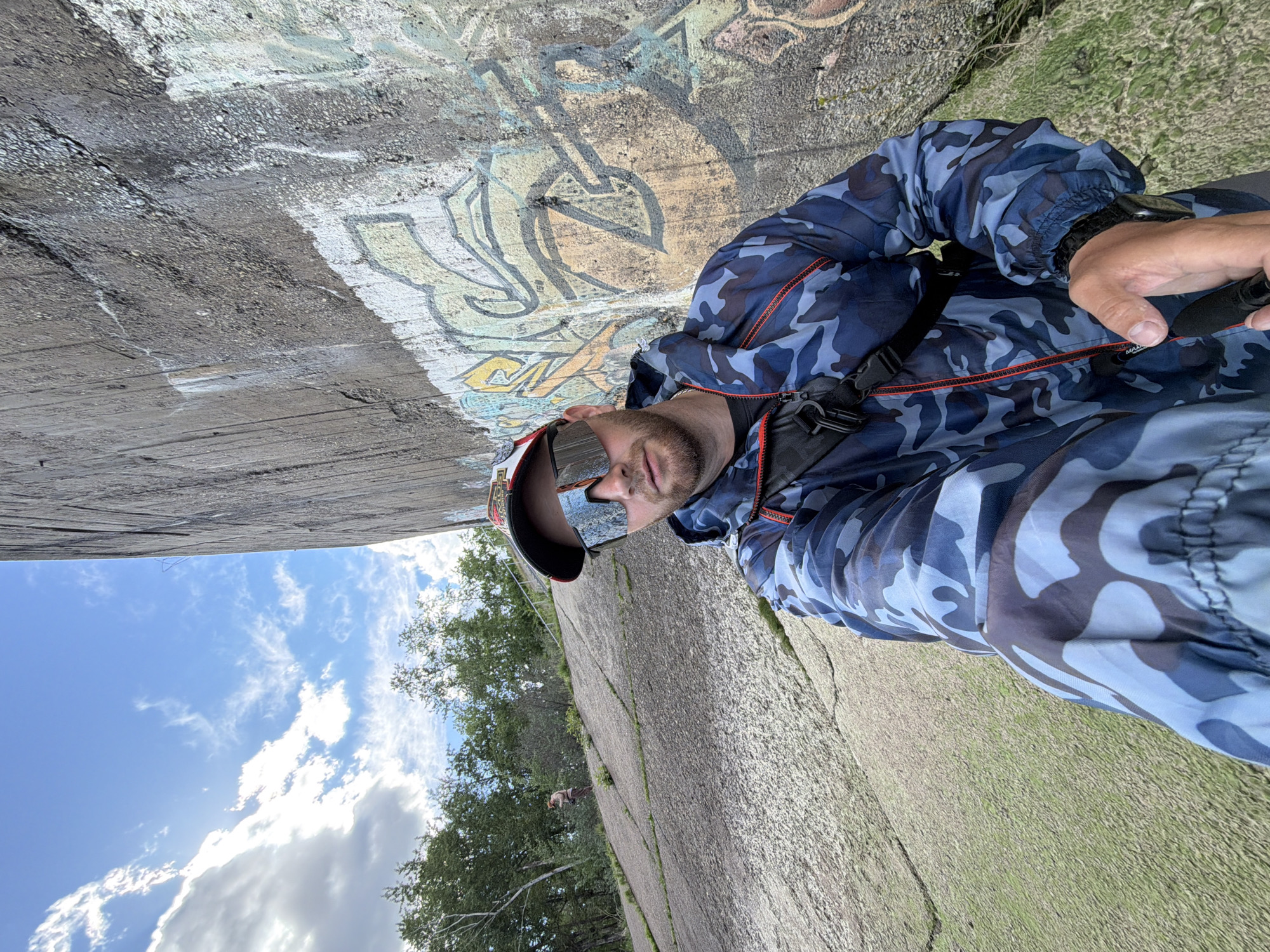 A man wearing a blue camouflage jacket and mirrored sunglasses stands near a concrete structure with graffiti. The location is Chaikovsky, Russia. The sky is visible in the background, with clouds. The ground is covered with vegetation.