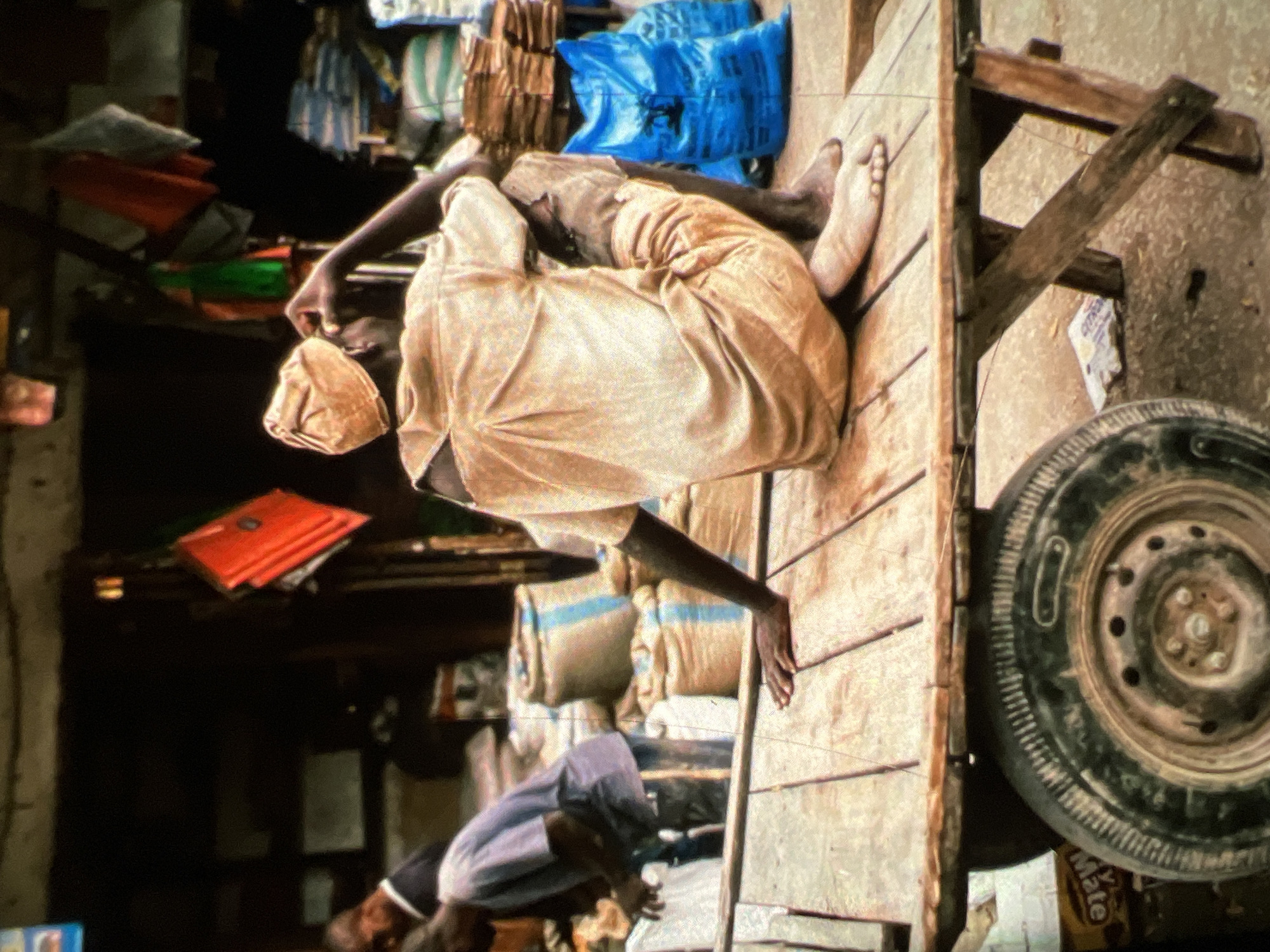 The image depicts a bustling market scene, likely in Lagos, Nigeria, characterized by its vibrant yet unorganized setting. In the foreground, a man wearing a light-colored tunic and cap is seated on a wooden cart. His posture suggests contemplation or rest, with one hand raised to his face and his legs crossed. The cart itself is rustic, with a visible tire and wooden planks forming its surface.
Behind the seated man, another individual in a dark t-shirt and light pants is seen tending to goods, possibly arranging or displaying them. The background is filled with various market elements. Stacks of goods, some wrapped in burlap sacks with blue stripes, are visible. Brightly colored items, like red bags or fabrics, are hanging, adding to the visual texture of the market. Large blue plastic bags are also present, hinting at merchandise or supplies. The overall environment appears to be an outdoor market, with a mix of natural light and the shadows cast by stalls and structures. The ground is uneven, composed of dirt and scattered debris, common in such informal trading spaces. The scene captures a moment of daily life within a Nigerian urban market, emphasizing the activity and the human element amidst the commercial setting.