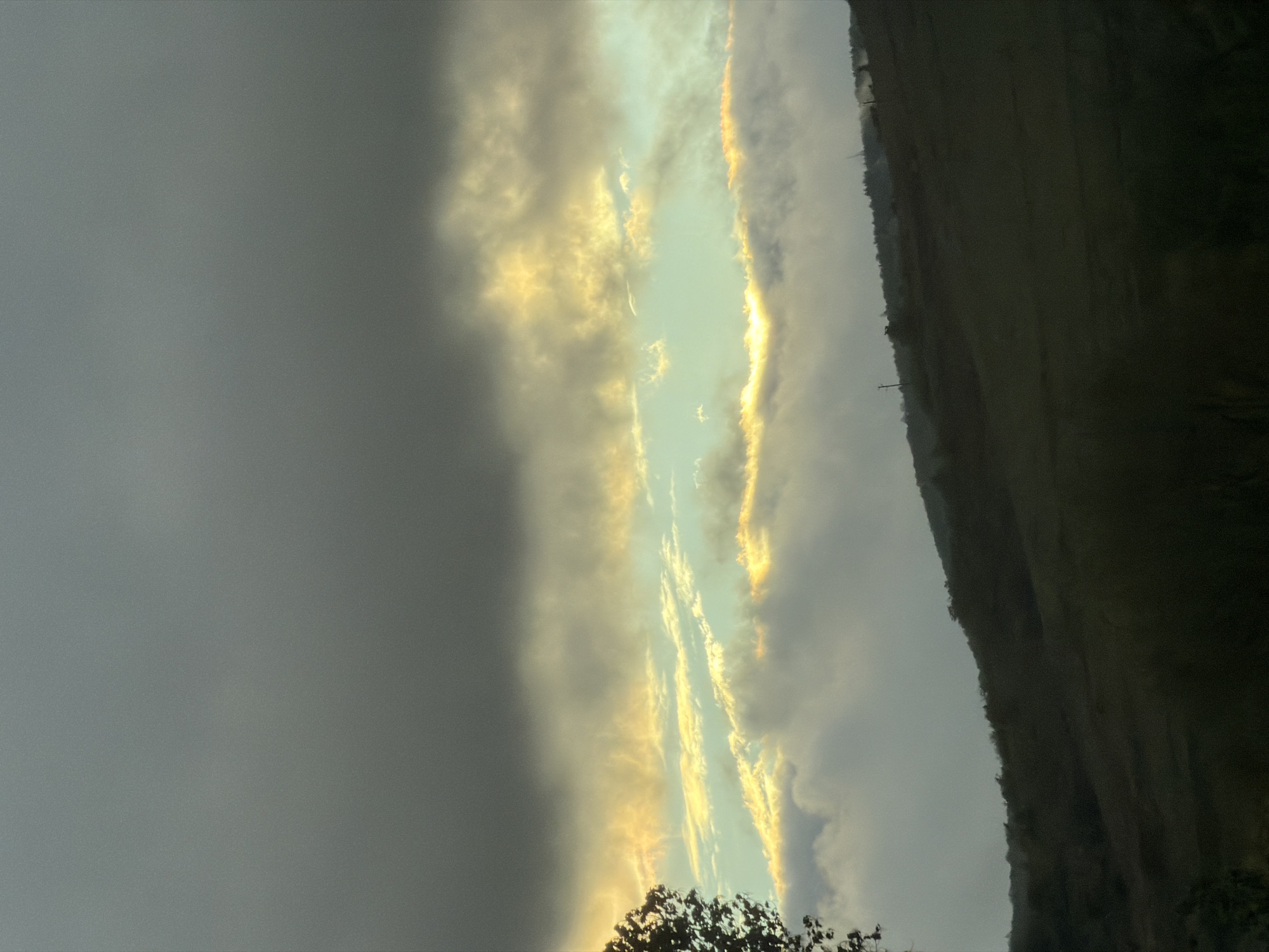 The image displays a dramatic and atmospheric natural landscape, likely captured during either sunrise or sunset, over Blajievo, Bulgaria. The dominant feature is the sky, divided into distinct sections by cloud cover and light.
On the left side, the sky is dominated by heavy, dark gray clouds, suggesting an overcast or stormy condition in that area. Contrasting sharply with this, a central vertical band of the sky is illuminated by intense, warm golden light, breaking through thinner, lighter clouds. This light creates striking streaks and patterns, highlighting the edges of the clouds with vibrant hues of yellow and orange. The area directly lit by the sun appears to be a light greenish-blue, indicating clear sky where the sun penetrates.
To the right, a dark, undulating landmass forms a prominent silhouette against the lighter sky. This appears to be a hill or mountain, covered densely with vegetation, primarily trees, which are indiscernible in detail due to the low light. A few distinct tree crowns are visible at the very bottom edge of the frame, appearing as dark shapes against the glowing sky. A faint, slender, pole-like structure can be observed on the distant hill, suggesting a human-made element.
The overall scene conveys a sense of natural grandeur and striking light conditions, characteristic of twilight hours when the sun is low on the horizon. The weather is partly cloudy, with a clear distinction between dense cloud cover and areas where sunlight is breaking through. There is no visible text in the image.