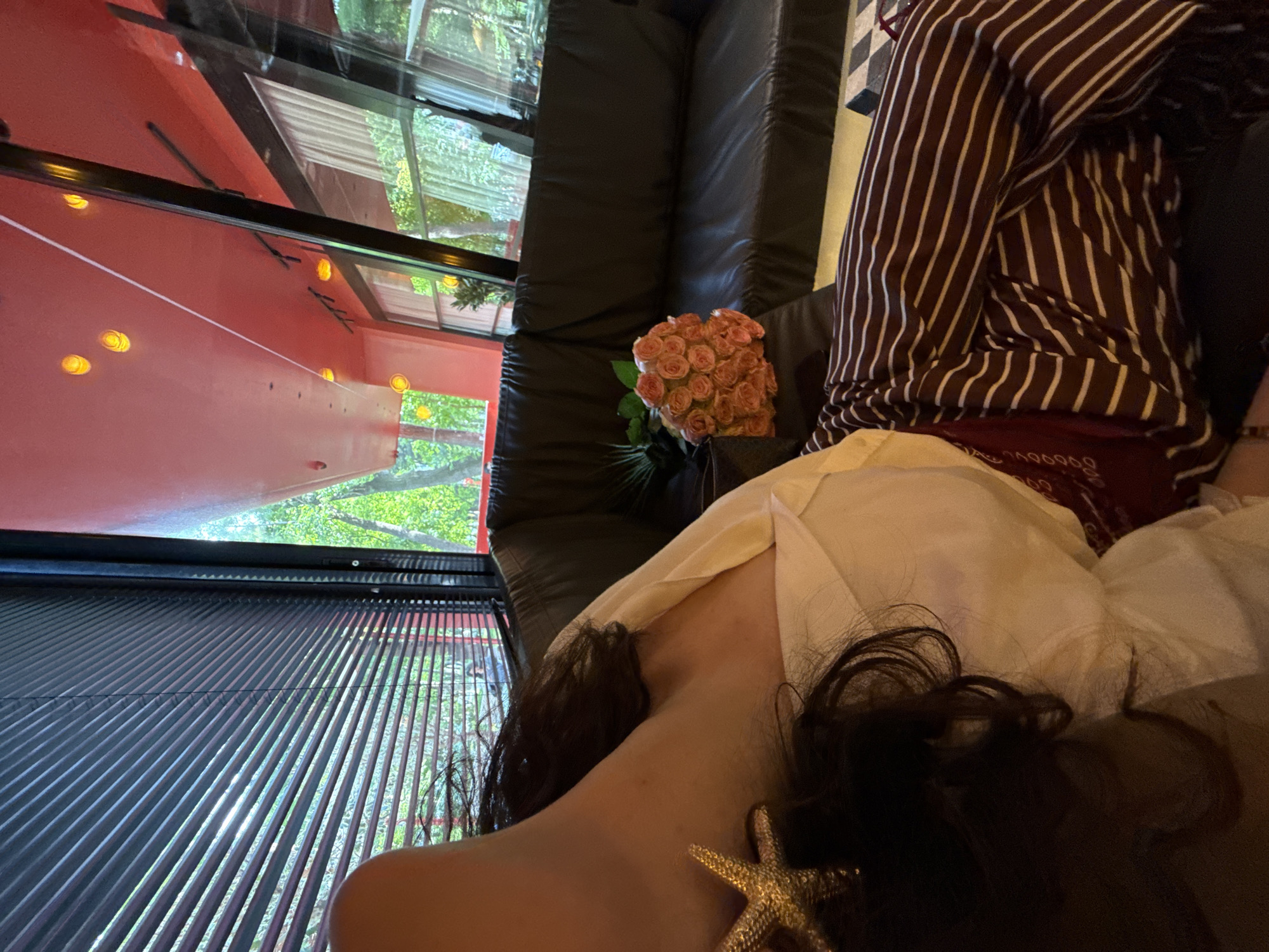 The image captures a scene inside a restaurant, possibly in Yerevan, Armenia, given the distinctive red-painted exterior visible through the windows. The foreground shows a person's bare shoulder, with dark, curly hair adorned with a gold starfish accessory. They are lying down, covered with a white cloth or sheet.
Next to the person is a bouquet of pale pink roses. The person in the photo is wearing a horizontally striped shirt. The seating area features a black, plush backrest. Outside the windows, greenery and natural light suggest a garden or park setting. The windows themselves have a slatted or partially obscured design.