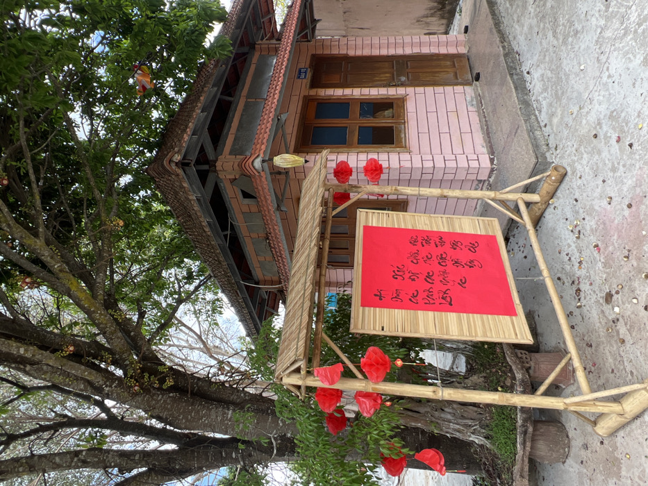 This image depicts an outdoor scene in Đăk Mar, Vietnam. The central foreground features a bamboo-framed display stand with a woven bamboo canopy. Mounted within this frame is a large, rectangular red board adorned with black Vietnamese calligraphy. Several red paper lanterns are suspended from the bamboo structure and from the branches of a large, leafy tree positioned to the left. The tree exhibits a thick, textured trunk and numerous globular fruits hanging directly from its branches. The base of the tree is contained within a circular, raised concrete planter. In the mid-ground, to the right, stands a small, single-story building constructed of light-pink bricks, featuring a multi-tiered, reddish-brown tiled roof. The building has a dark wooden door on its right side and two dark-paned wooden-framed windows on its front face. A small sign with "BẢO VỆ" (Vietnamese for "Guard") is affixed above the door. A yellow ornamental lantern hangs from the building's front corner. The ground surface is paved with grey concrete, upon which numerous small, round, light-colored objects, possibly fallen fruit or seeds, are scattered. The sky overhead is partially visible, appearing light blue. No individuals are present in the scene.