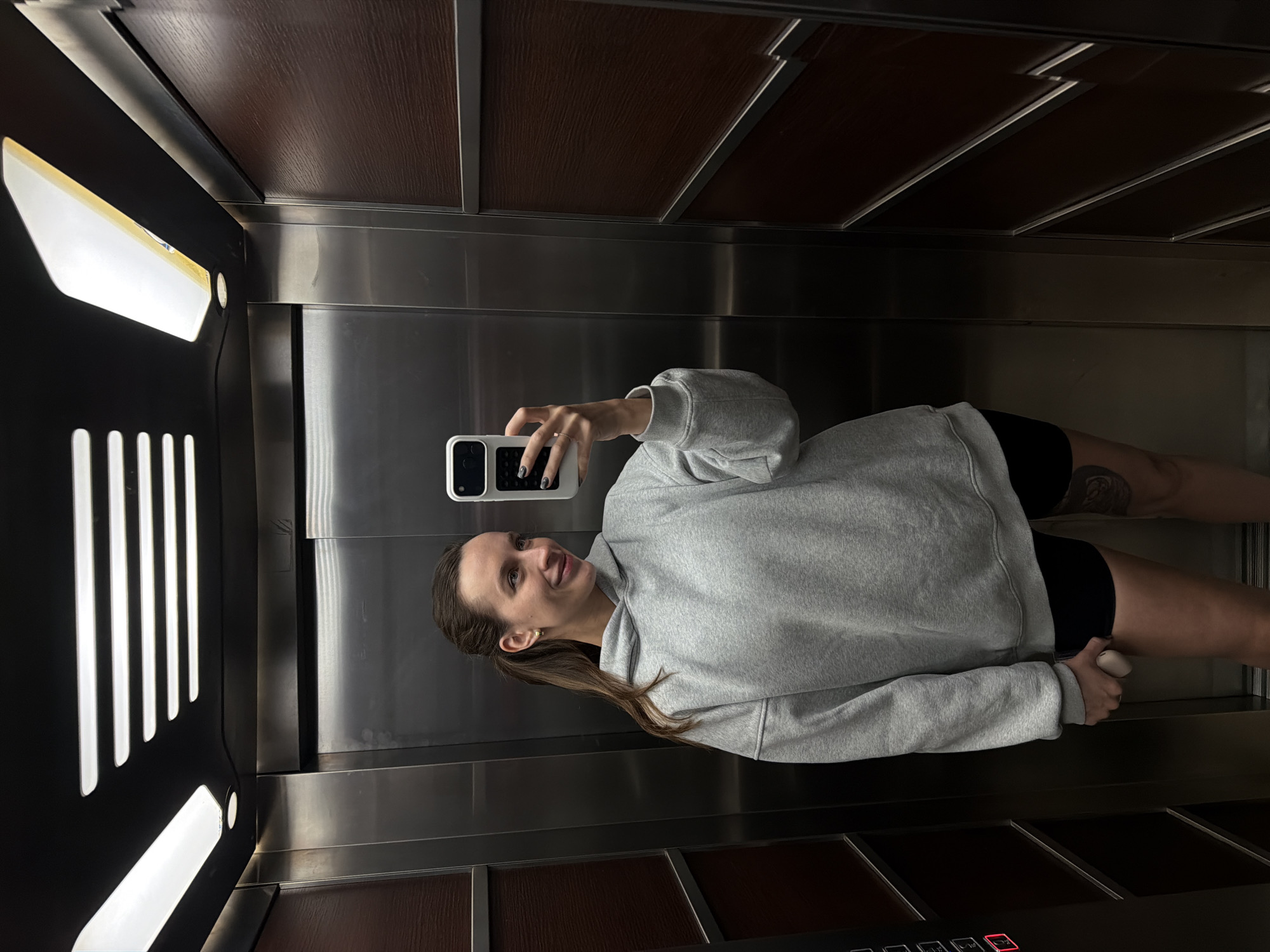 A woman is taking a selfie in an elevator. The elevator has wooden paneling, stainless steel walls, and elongated vertical lights. The woman is wearing a gray sweatshirt and black shorts. Her hair is in a ponytail. The exact location in Budva, Montenegro cannot be determined from this image.