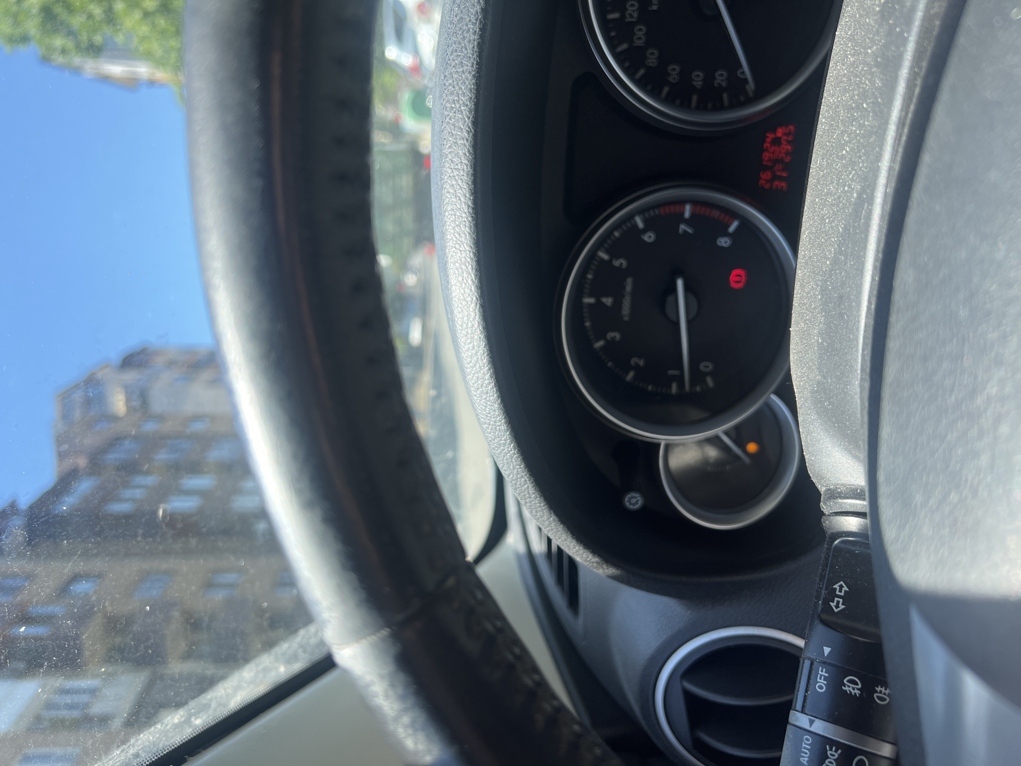 The image shows a partial view from inside a car. The steering wheel is on the left, and the dashboard instruments, including a tachometer showing approximately 1,200 RPM and a speedometer, are visible to the right. The GPS coordinates "26.1924 E, 42.4975 N" are displayed on the dashboard. These coordinates locate the position in Burgas, Bulgaria. A building is visible through the windshield.