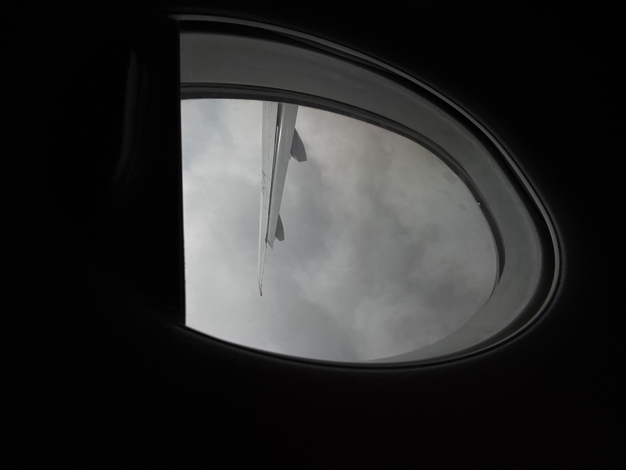 The image shows a view from an airplane window looking out at a cloudy sky and a section of an airplane wing. The foreground is dark, obscuring the interior of the plane, and frames the view of the wing and sky. The wing appears to be white with some red markings near the tip. The sky is filled with dense, gray clouds, suggesting overcast weather. There are no people or identifiable landmarks visible that would indicate the specific location in Staroe Mart'yanovo, Russia. The image captures a moment of flight, characterized by the limited visibility and the presence of the aircraft's wing as the primary subject outside the window.