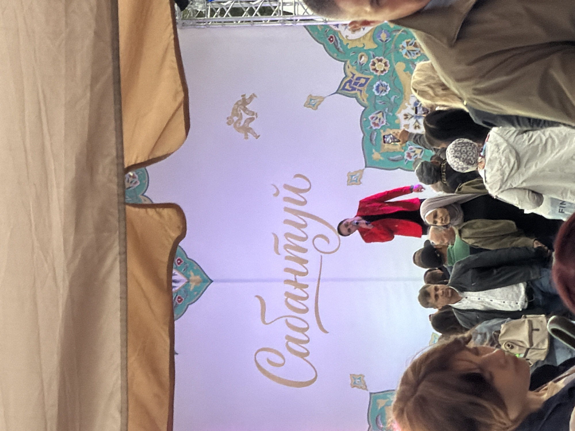 A man in a red jacket and dark shirt stands on a stage in front of a white backdrop with intricate designs and text that reads "Сабантуй". He is holding a microphone and appears to be speaking. A crowd of people are gathered in front of the stage, some looking towards the performer, while others are looking at their phones or talking amongst themselves. The scene appears to be an outdoor event, possibly a festival, as indicated by the stage setup and the presence of a large tent or awning above. The overall atmosphere is one of a public gathering or performance.