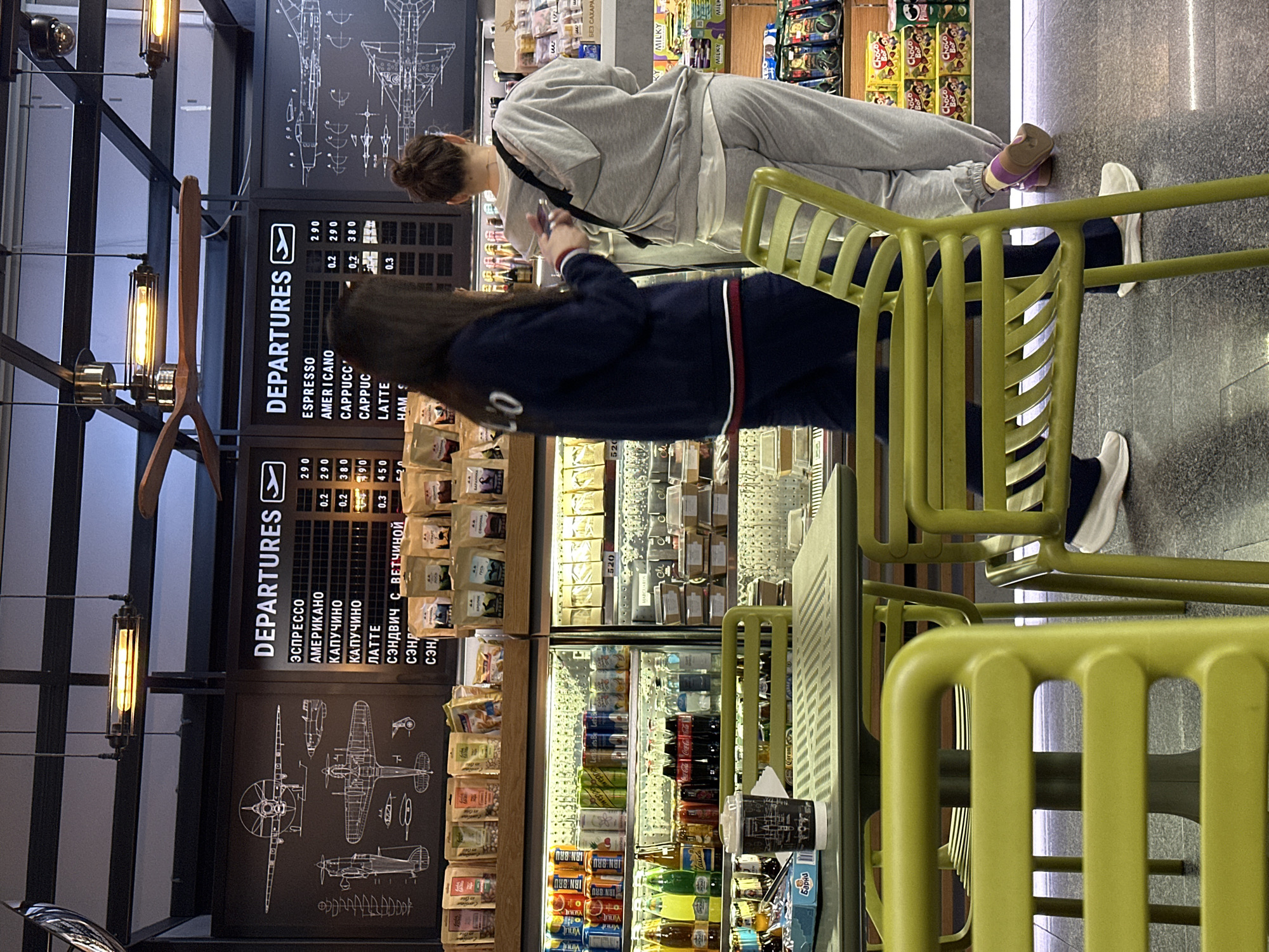 A retail space, possibly a café or convenience store, displays a blackboard with the word "DEPARTURES" in Cyrillic and English, alongside a price list for espresso, americano, cappuccino, and latte. Two female customers are visible; one is wearing a gray tracksuit and the other is wearing a dark blue jacket with a white numeral. They are standing near shelves stocked with a variety of food and beverage items, including snacks, bottled drinks, and coffee. The café furniture includes green chairs.
The location is likely in Sochi, Russia. This is determined by the use of Russian Cyrillic script on the Departures board.