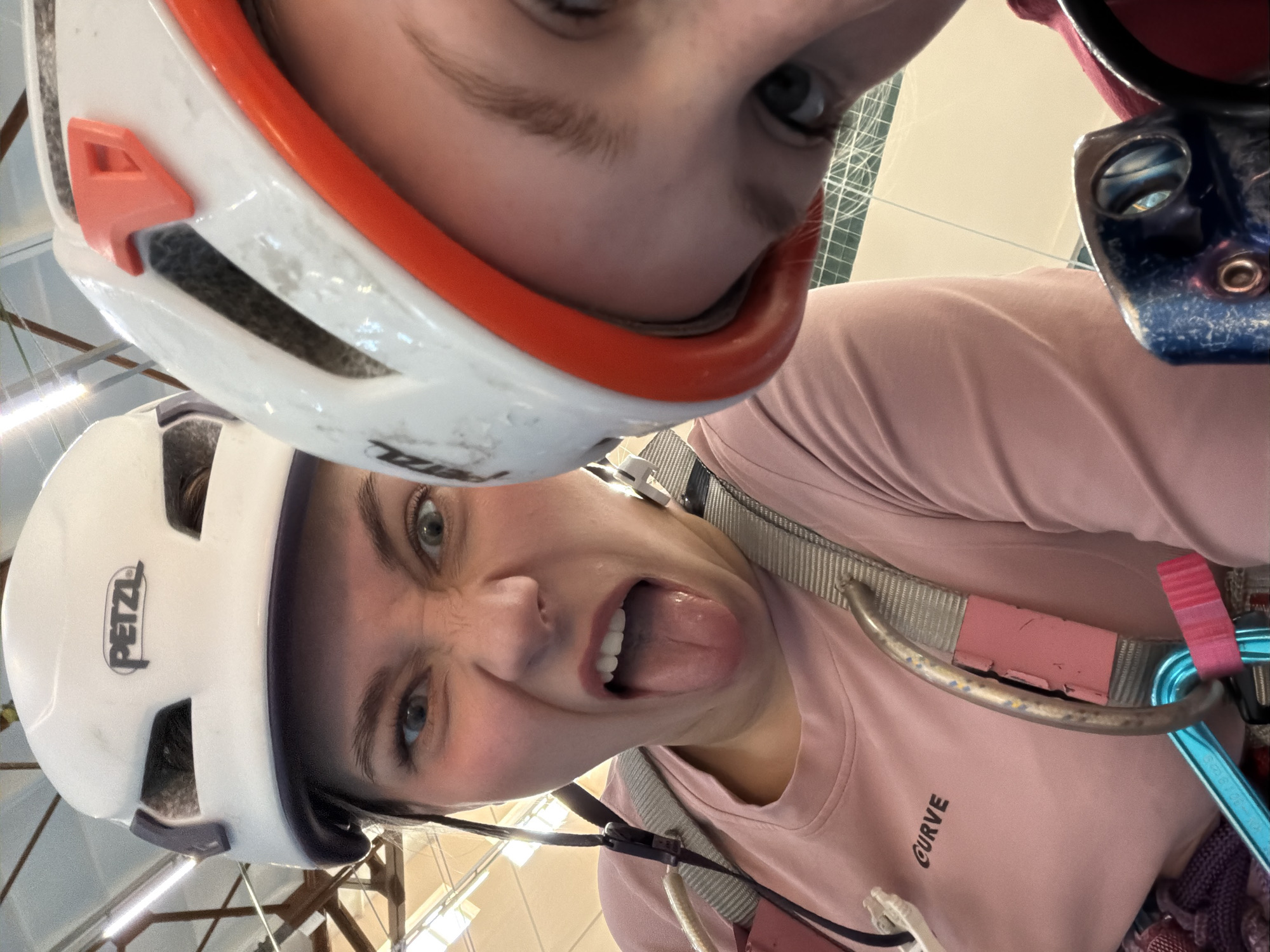 The image shows two people posing closely for a selfie-style photograph. Both are wearing climbing helmets. The person in the foreground is wearing a white Petzl helmet and a pink t-shirt with the word "CURVE" printed on it. They are sticking their tongue out and making a silly face. They also appear to be wearing climbing gear, including a harness and carabiners. The person in the background is also wearing a climbing helmet, white with an orange brim. The location appears to be indoors, possibly a climbing gym, given the presence of climbing equipment and the overhead lighting and structural beams. The image does not provide enough detail to confirm a specific location in Berezniki, Russia.