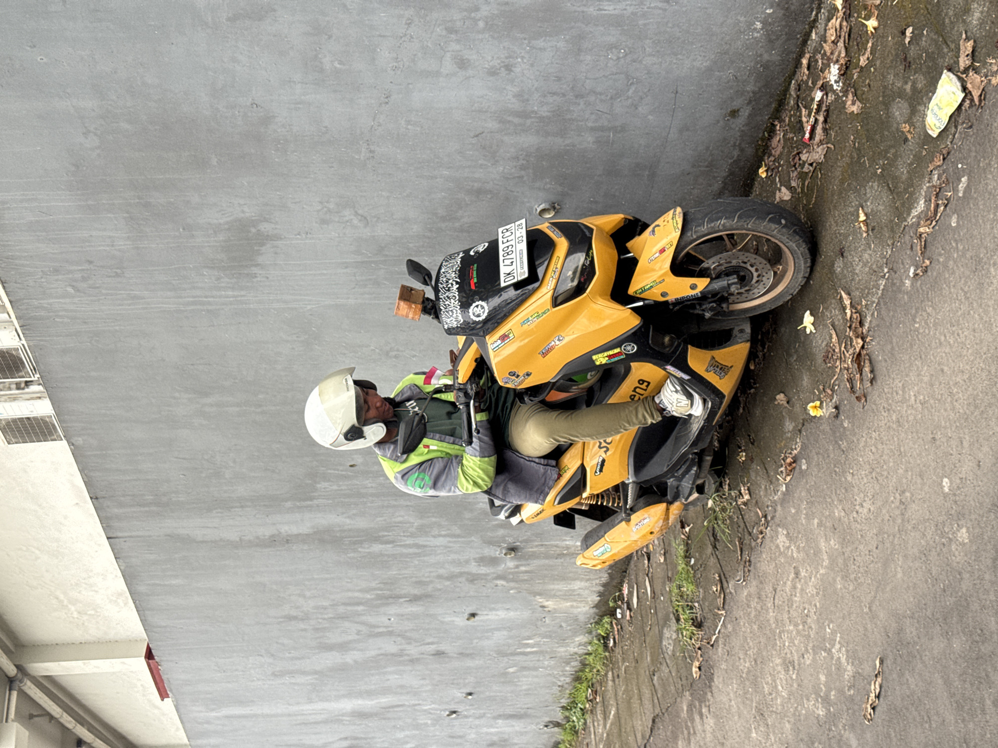 The image, rotated 90 degrees clockwise, captures a man sitting on a bright yellow motorcycle in what appears to be an outdoor urban or semi-urban setting. The scene is set against a plain, tall grey concrete wall on the left, with a narrow strip of paved ground and accumulated debris (leaves, dirt, and some trash) along the base of the wall on the right.
The central subject is a man wearing a white full-face helmet and a distinctive green and grey hi-visibility vest over a dark long-sleeved shirt. He also wears light-colored pants and white sneakers. The man is positioned on the yellow scooter, holding a smartphone with both hands and looking down at it, suggesting he is either using it or waiting.
The motorcycle is predominantly yellow with black accents and numerous stickers covering various parts of its body. A rectangular wooden block is conspicuously attached to the left handlebar. The front license plate is clearly visible, displaying "DK 4789 FCR" and "03 - 28" below it, indicating it's from Indonesia. Other visible text includes "YAMAHA" and parts of other company logos or slogans on the bike's stickers, though most are too small to read.
The ground next to the motorcycle is concrete or asphalt, scattered with dried leaves and some light green weeds growing in cracks. An empty, crumpled plastic wrapper is visible among the debris. In the top left corner, part of a building with vents is visible. The lighting suggests it is daytime. The context indicates the location is Kuta Selatan, Indonesia.