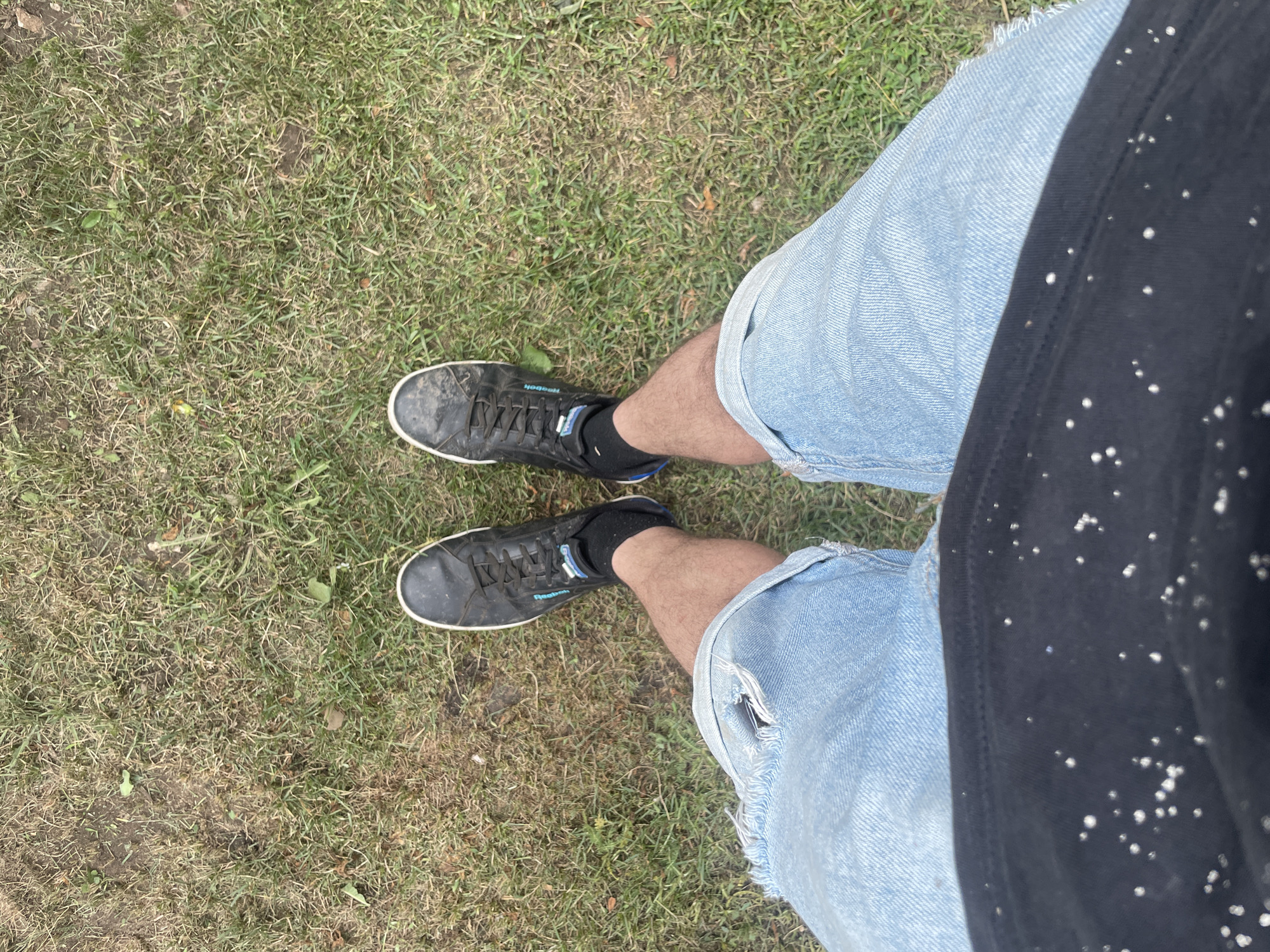 This is a first-person perspective, eye-level medium shot of a person's legs and feet standing on a grassy area. The person is wearing distressed light-wash denim shorts and black sneakers with white soles and black socks. The grass is a mix of green and dry patches, with some scattered leaves. The background consists solely of this grassy terrain. The image was captured on August 14, 2023, at 16:14:37 in Laishevo, Russia.