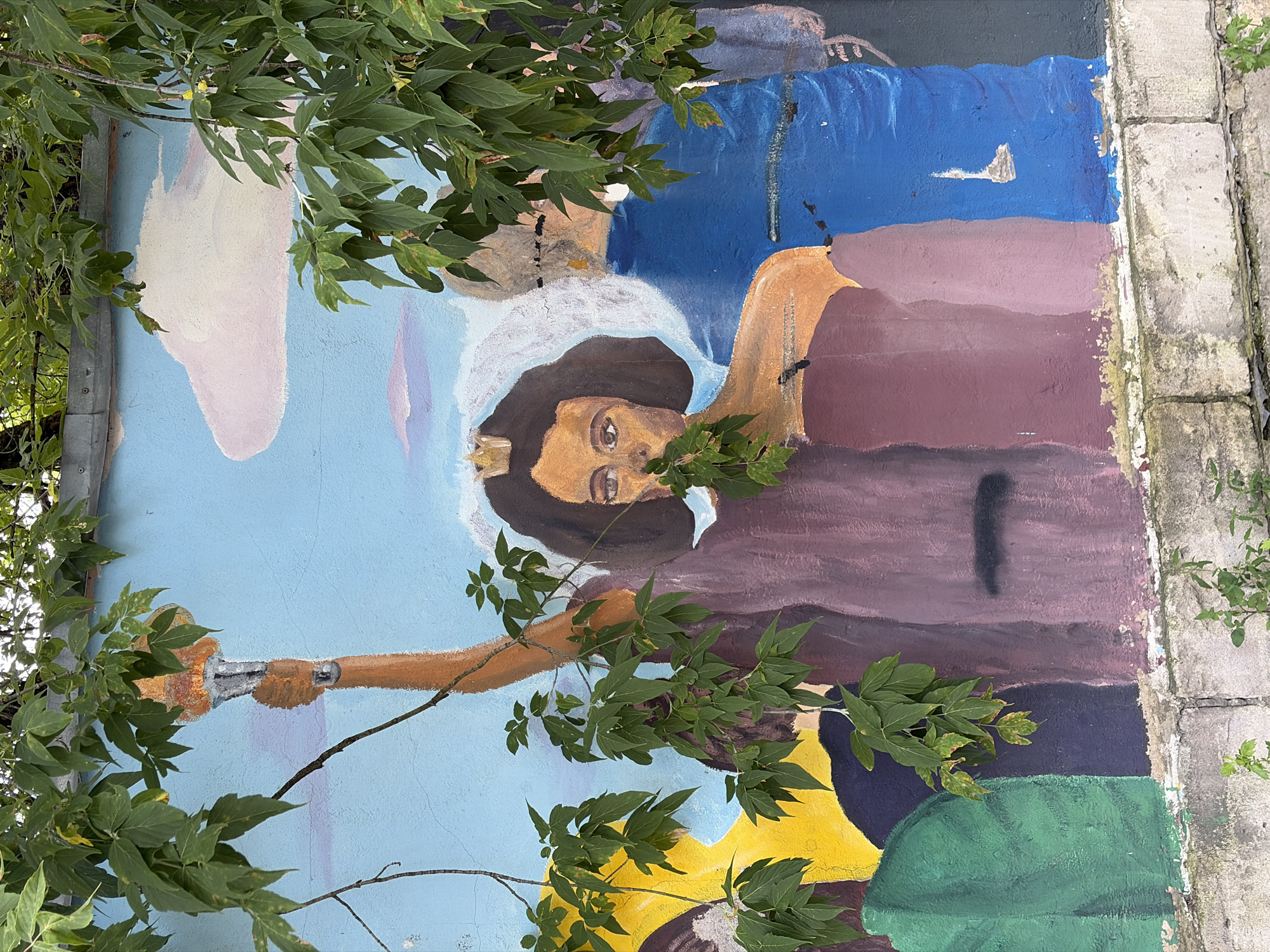 This is a photograph of a mural on a wall, partially obscured by green foliage. The mural depicts several figures in a stylized manner. The central figure is a woman with dark, curly hair wearing a golden crown, her face turned slightly towards the viewer, and her right arm raised holding a torch. She is adorned in a purple garment. To her left, a portion of another figure in yellow is visible. To her right, another figure in a bright blue garment is partially depicted. The background of the mural features a light blue sky with soft pink and white clouds. The mural is painted on a textured, concrete wall, which shows signs of wear and tear, with visible cracks and chipping paint. The base of the wall is constructed from rough, grey concrete blocks. The foliage in the foreground consists of green leaves and thin branches. This mural is located on the exterior wall of a building in the city of Khimki, Russia.