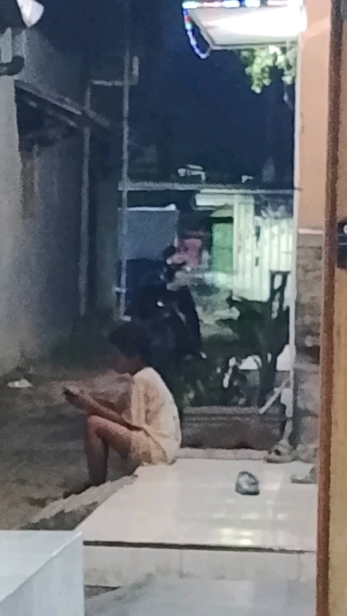 A person is seated on a step in an alleyway, looking down at a mobile device. The person is wearing a light-colored shirt and appears to be crouching. The alleyway is paved with light-colored tiles, and there are various objects and structures visible in the background, including a building with a green door, potted plants, and what appears to be a motorcycle. The lighting suggests it is nighttime or dusk. The overall scene is dimly lit, with some artificial light sources creating reflections on the tiled surface.