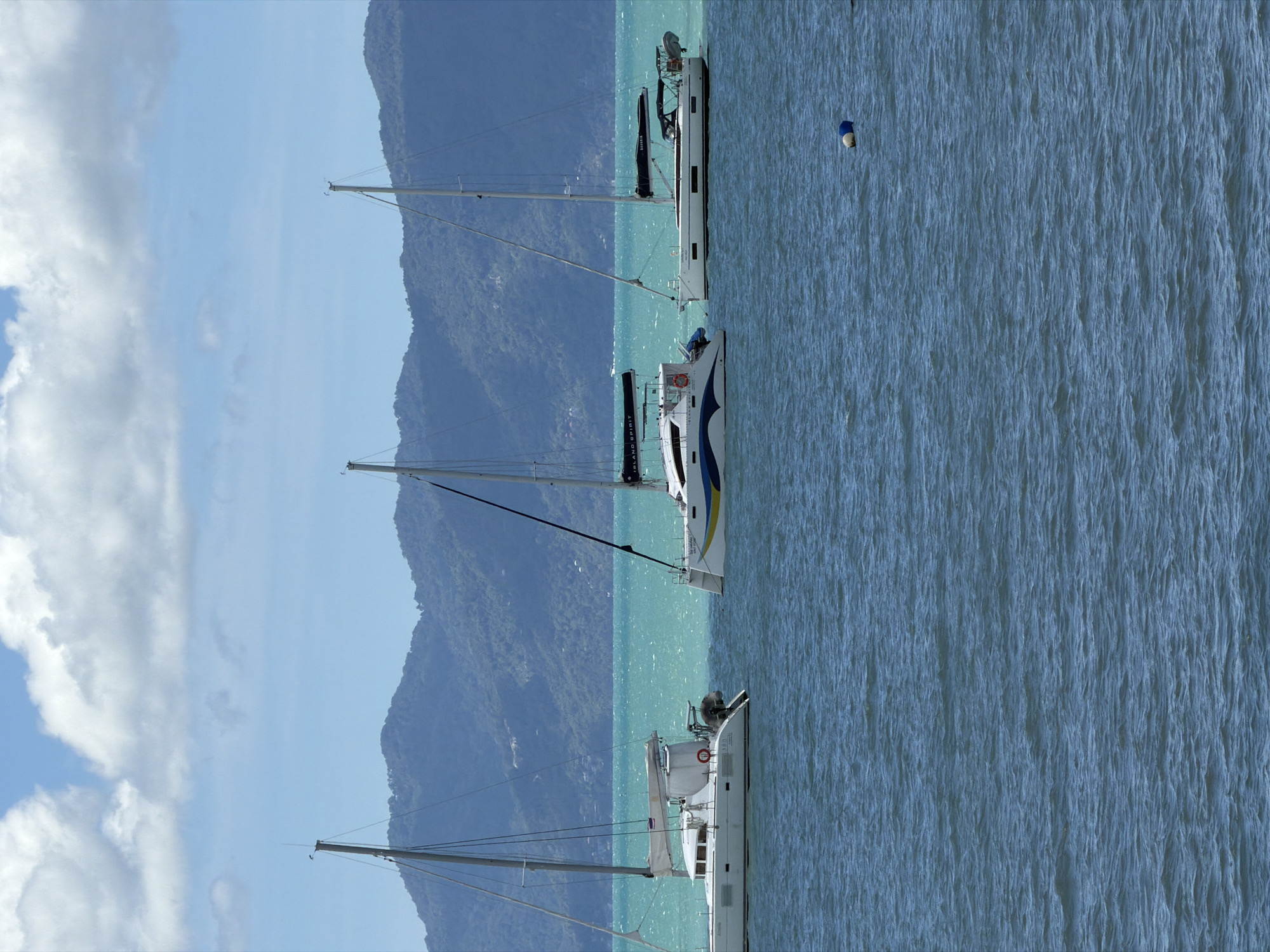 Three white sailboats are moored in the ocean off the coast of Baan Bo Phut, Thailand. The water is a vibrant turquoise, and lush green mountains form the backdrop. The sky is a brilliant blue, dotted with fluffy white clouds. The sailboats are modern and sleek, with tall masts and white sails. One boat, a catamaran, is on the left, and two monohull sailboats are to its right. The water is choppy, suggesting a light breeze. A small blue and white buoy floats in the water in the foreground. The scene is peaceful and idyllic, a perfect representation of a tropical paradise.