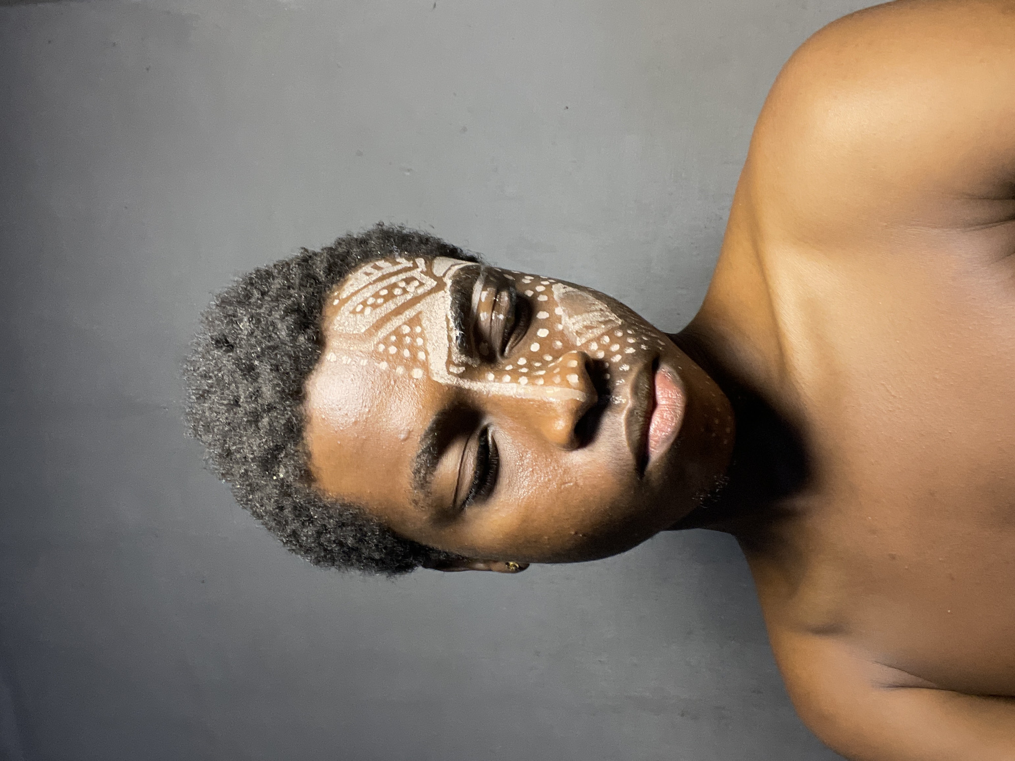 The image shows a person with dark skin, dark hair, and closed eyes, with white paint designs on their face. The person is shirtless and appears to be lying down against a grey backdrop. The environment appears to be indoors. There is no visible indication that this image was taken in Nkpor, Nigeria.
