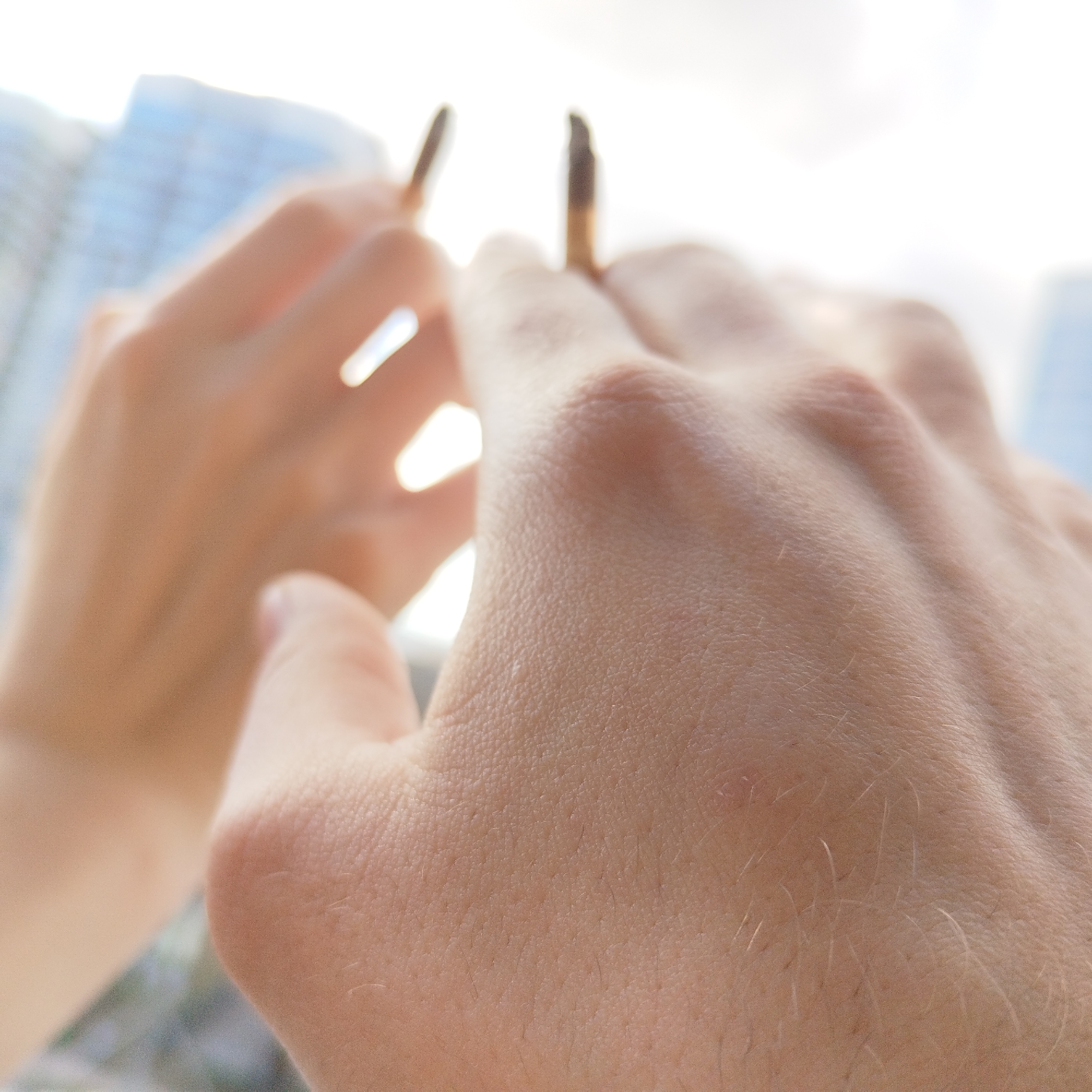 The image shows two hands holding two objects that appear to be chopsticks. The location is suggested to be Yekaterinburg, Russia, though no specific identifiable elements of the city are visible within the frame. The background includes blurred outlines of buildings, hinting at an urban environment. The focus is on the texture and details of the skin on the hands.