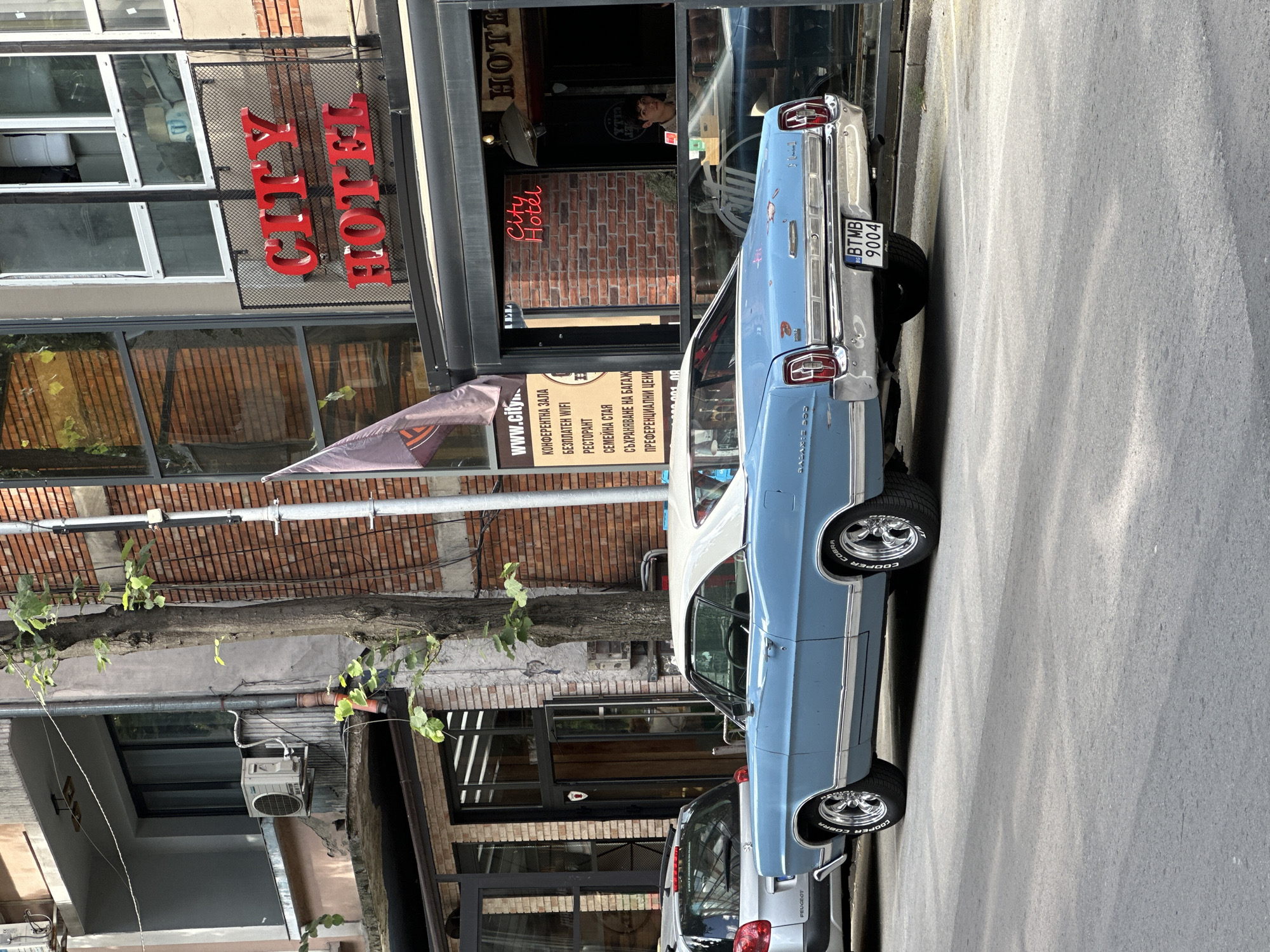 The image shows a light blue vintage car parked on a street in front of a building. The building appears to be a hotel, with a prominent red sign reading "CITY HOTEL" in large letters. Another sign, also in red neon, inside a window also says "City Hotel." There are various signs on the building with text in Bulgarian, indicating services like "CONFERENCE HALL," "FREE WIFI," "RESTAURANT," and "FAMILY ROOM." A person can be seen through a window inside the hotel. The car has a license plate that reads "BTMB 9004" and the word "PEUGEOT" is visible on the back of another car parked behind it. The tires of the vintage car have "COOPER COBRA" written on them. The scene suggests a sunny day, as indicated by the shadows cast on the street. The location is identified as Veliko Tarnovo, Bulgaria. The overall impression is one of a street scene featuring classic automotive design against a backdrop of local businesses.
