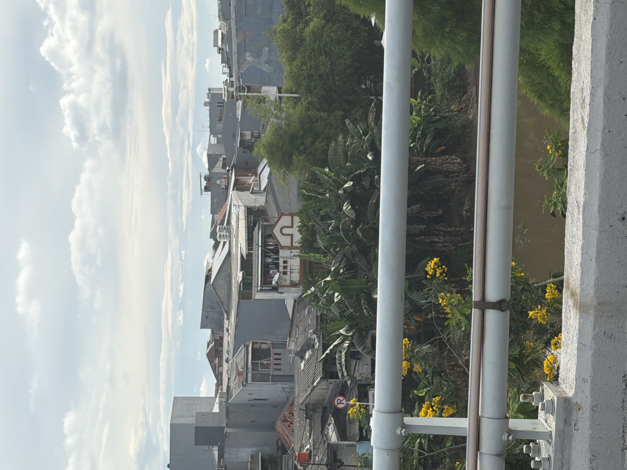 The image captures a scene in North Jakarta, Indonesia, overlooking a dense urban environment. The foreground is dominated by a bridge railing, with yellow flowering plants intertwined. Beyond the railing, there's a view of closely packed buildings, a mix of residential and commercial structures, typical of densely populated areas. Banana trees and other greenery add to the cluttered landscape. The sky is partly cloudy, suggesting a humid tropical climate. The architecture is varied, with visible satellite dishes on some rooftops, indicating modern amenities amidst older constructions.