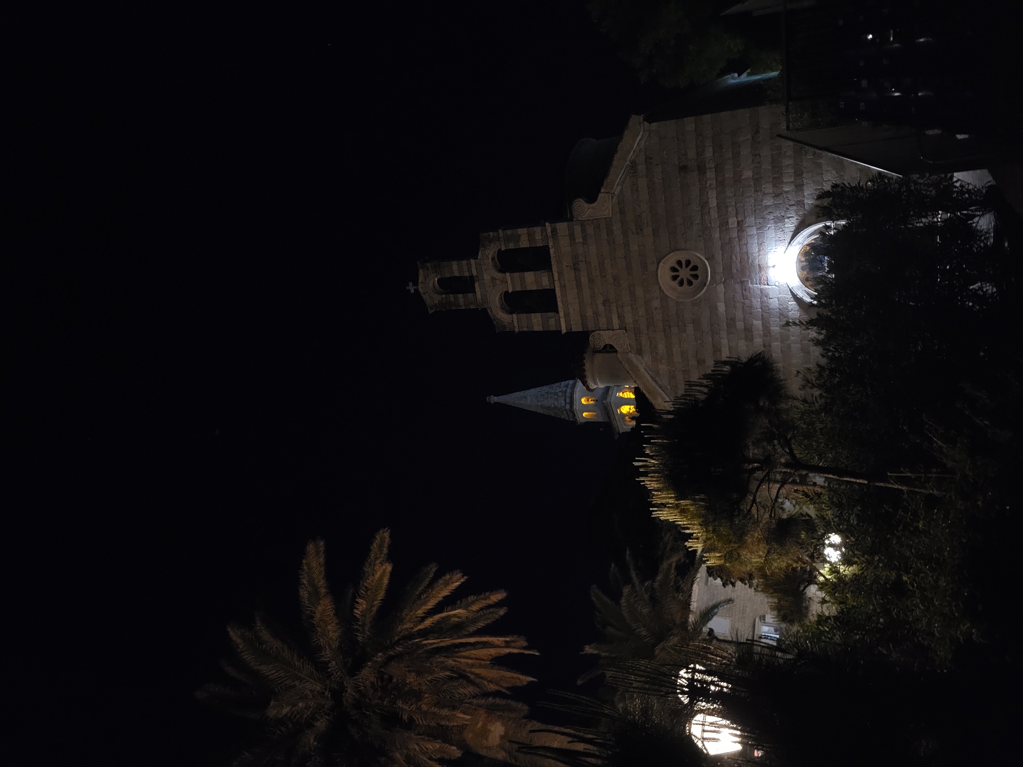 Captured at night, the image shows a church in Budva, Montenegro. The building features light-colored stone construction, a bell tower with arched openings, a round window, and a spire-like tower with glowing lights within. Palm trees are in the foreground. Other vegetation, including trees, surrounds the church. The background is a dark night sky.