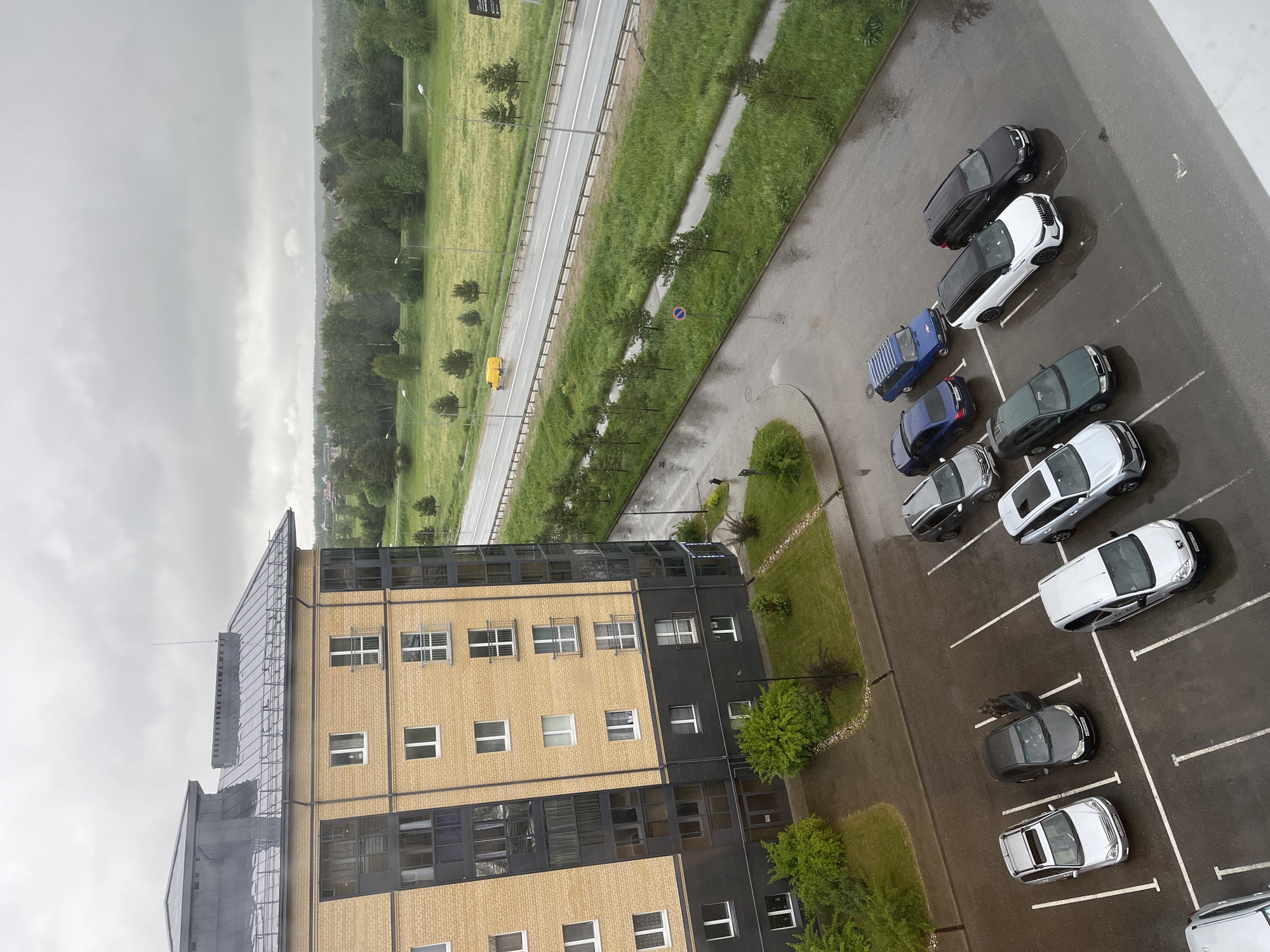 The image depicts an aerial view of an urban landscape, likely in Mokshino, Russia, characterized by a mix of infrastructure and natural elements under an overcast sky. The foreground is dominated by a multi-story building with a facade that combines yellow brick and black panels, punctuated by windows. To the right of the building is a parking lot with several parked cars of varying colors, including white, black, and blue. A patch of greenery with small trees and bushes separates the building and the parking lot. Beyond the parking lot, a multi-lane road runs parallel to a wide grassy field bordered by trees. A yellow van is visible on the road. The overcast sky suggests rainy or dull weather.