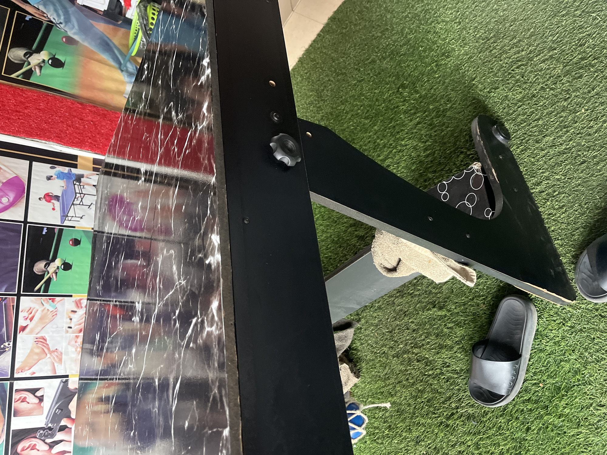The image shows a close-up of a black table with a marble-like surface. The table is supported by black legs that rest on a green artificial turf surface. A black flip-flop is visible on the turf to the left of the table, and another black flip-flop is partially visible at the bottom edge of the frame. A beige towel is draped over one of the table's legs. In the background, behind the table, are several framed pictures depicting various activities, including people playing billiards, table tennis, and what appears to be a beauty salon. The overall setting suggests an indoor recreational area, possibly a game room or entertainment center in Jalingo, Nigeria.