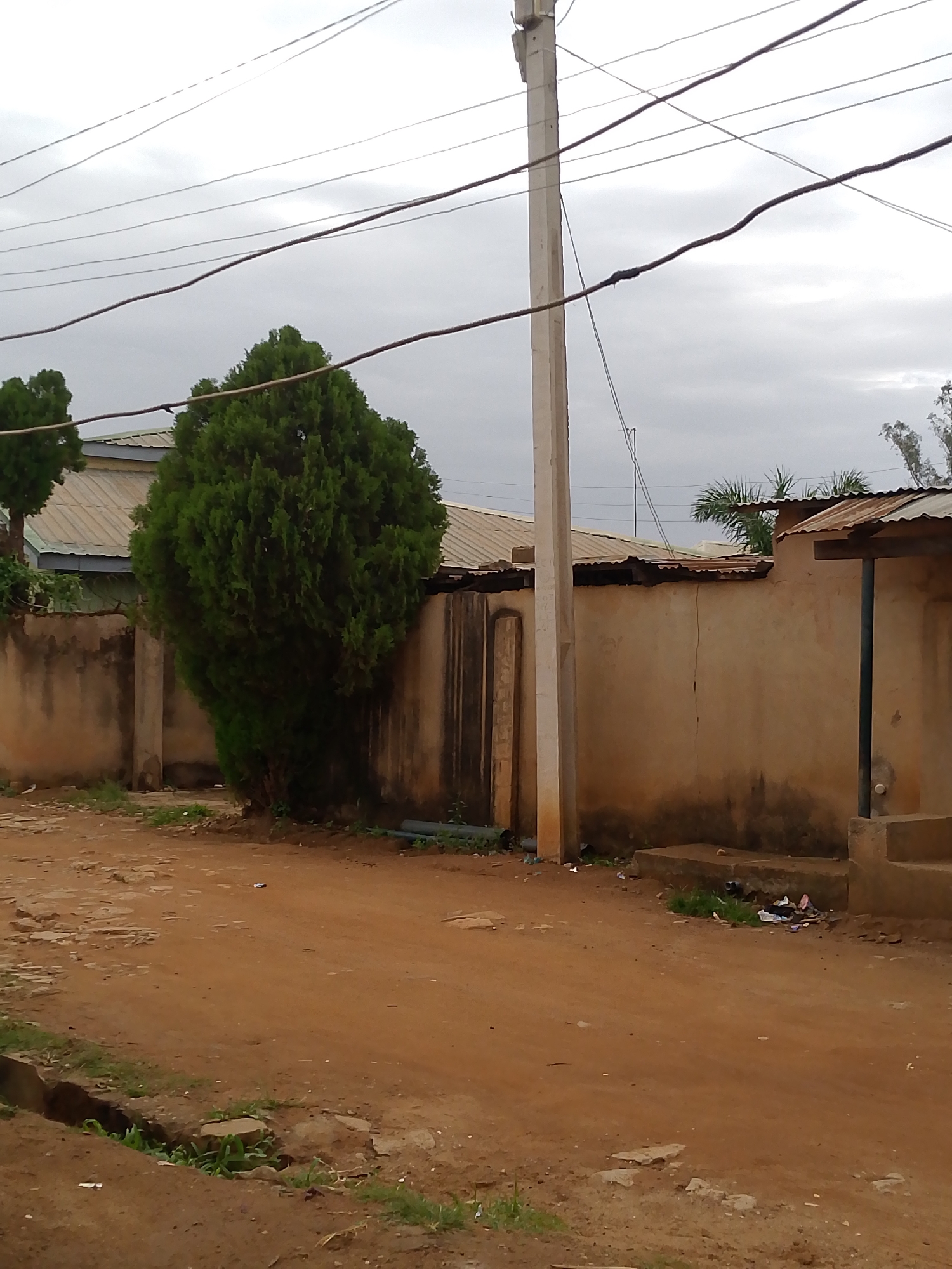 The image shows a street scene in Jos, Nigeria. The ground is red dirt. A wall runs along the right side of the street, constructed of stucco or similar material, and is dark-stained at the bottom. There is a covered section with a post supporting the overhang. A concrete step structure leads up to a doorway. A tree stands along the left side of the street against a similar concrete wall. Utility poles and wires are overhead. The sky is overcast. There is debris on the side of the street near the wall.