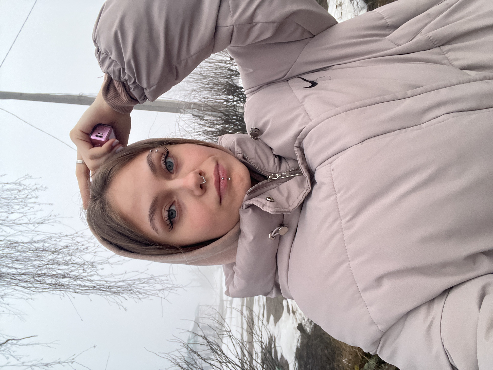 A young woman is pictured in a selfie, wearing a light purple puffer jacket. She has a nose ring and lip piercings. She is holding a small purple cube in her hand. The background appears to be a winter scene with bare trees and a misty landscape. The location is Фоки, Russia.