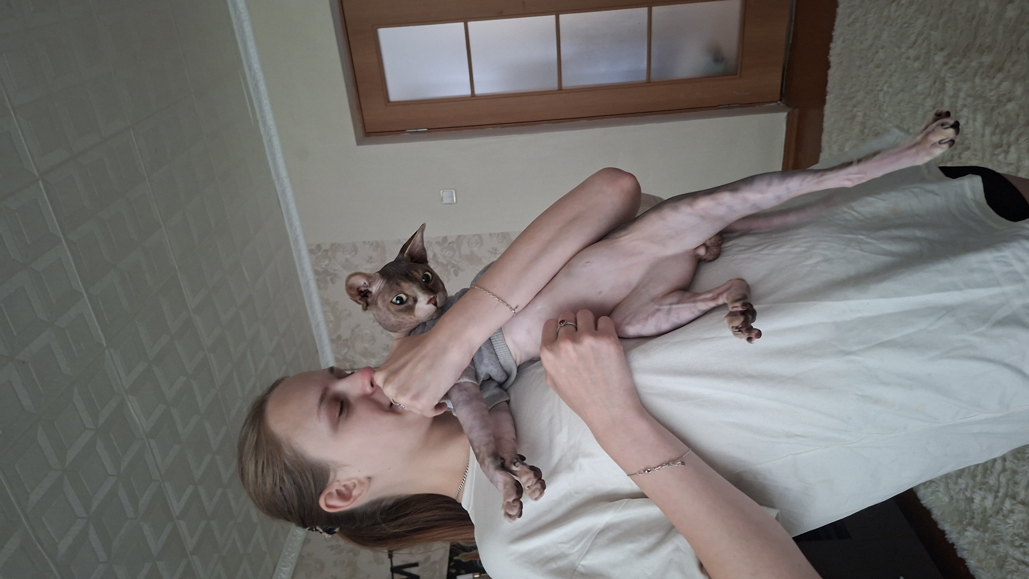 In a brightly lit, domestic interior that appears to be an apartment in Myadzyel, Belarus, a young woman with light brown hair pulled back in a ponytail is holding a hairless Sphynx cat. The woman is wearing a loose-fitting white t-shirt and is looking down at the cat, her hand near its face, suggesting a moment of affection or interaction. The cat, with its distinctive large ears and wrinkly skin, is wearing a grey sweater and is held securely against the woman's chest. The background shows a textured ceiling, wallpaper with a patterned design, and a closed wooden door with glass panes. A white rug is visible on the floor. The overall impression is one of a casual, everyday scene within a home environment.