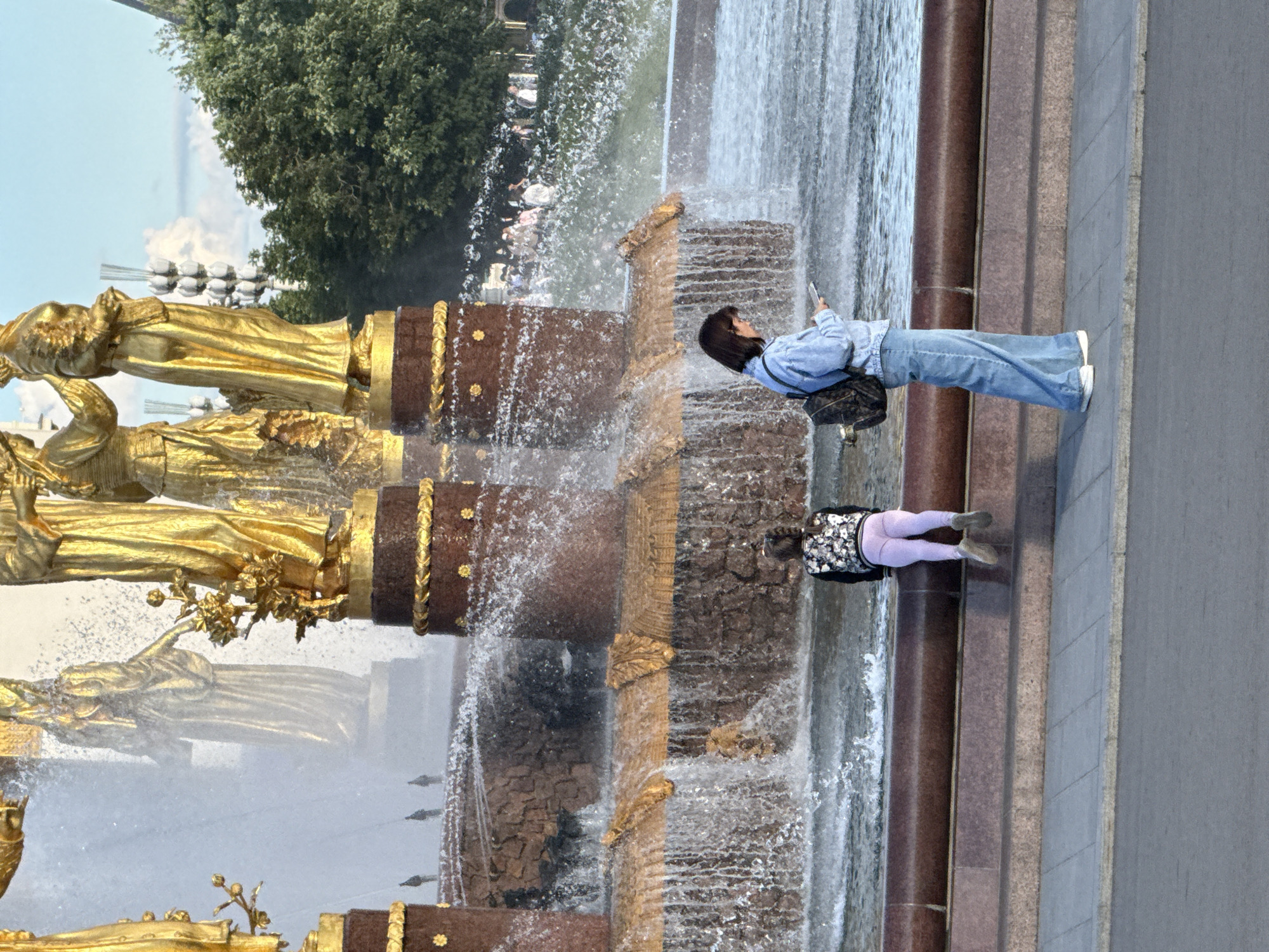 The image shows a section of the Friendship of Nations Fountain at VDNKh (Exhibition of Achievements of National Economy) in Moscow, Russia. In the foreground, two individuals are positioned near the fountain's cascading water. On the left, a young girl with braided hair is seated, facing away from the camera and towards the fountain. She is wearing a dark jacket with a floral pattern and light purple leggings. To the right, an adult, likely a woman, stands facing the fountain and looking down at a mobile device she holds in her hand. She is dressed in a light blue denim jacket, wide-leg blue jeans, and white sneakers, with a backpack slung over her shoulder. The fountain itself is grand, with golden statues of figures representing different republics of the Soviet Union visible in the background, partially obscured by water spray. The architectural elements of the fountain are ornate, featuring detailed carvings and gilded accents. The surrounding environment includes lush green trees in the background, suggesting a park or outdoor exhibition space. The sky is visible above, with a few clouds. The ground in the immediate foreground is paved with gray tiles, leading to a darker paved area, possibly a road. The overall scene captures a moment of leisure and observation at a prominent Moscow landmark.
