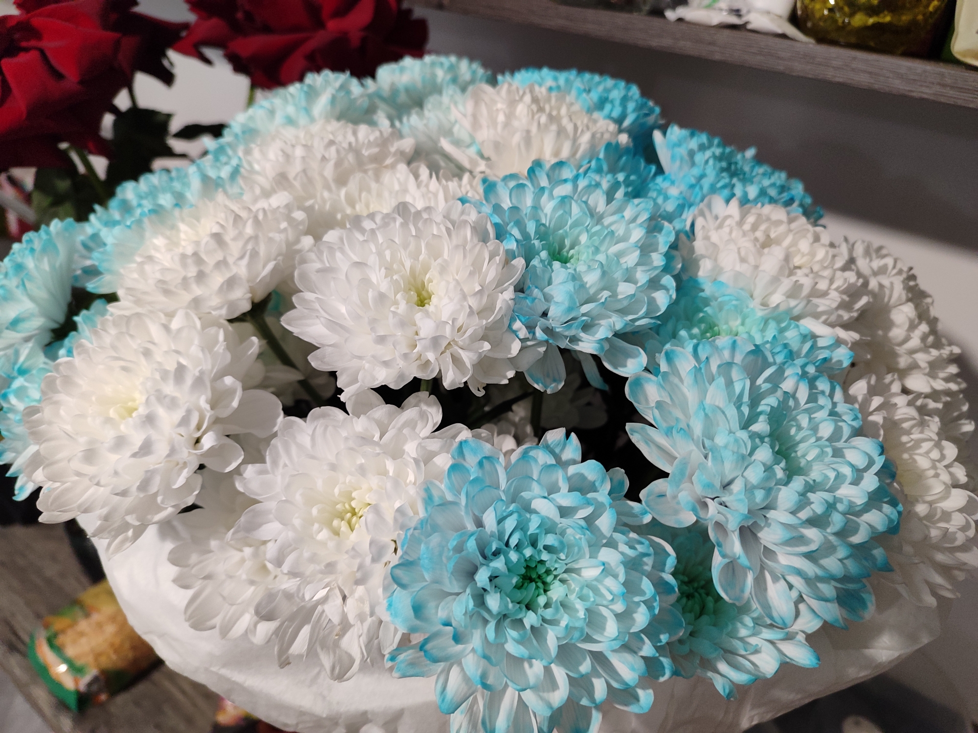 The image shows a bouquet of chrysanthemums, a mix of white and light blue varieties, arranged in what appears to be a white paper wrap. In the background, partially visible, are some red roses. The setting is likely indoors, with a shelf visible in the upper part of the image. There is no definitive information in the image to determine if it is in the city of Izhevsk, Russia. The image mainly features flowers and the immediate surrounding objects.