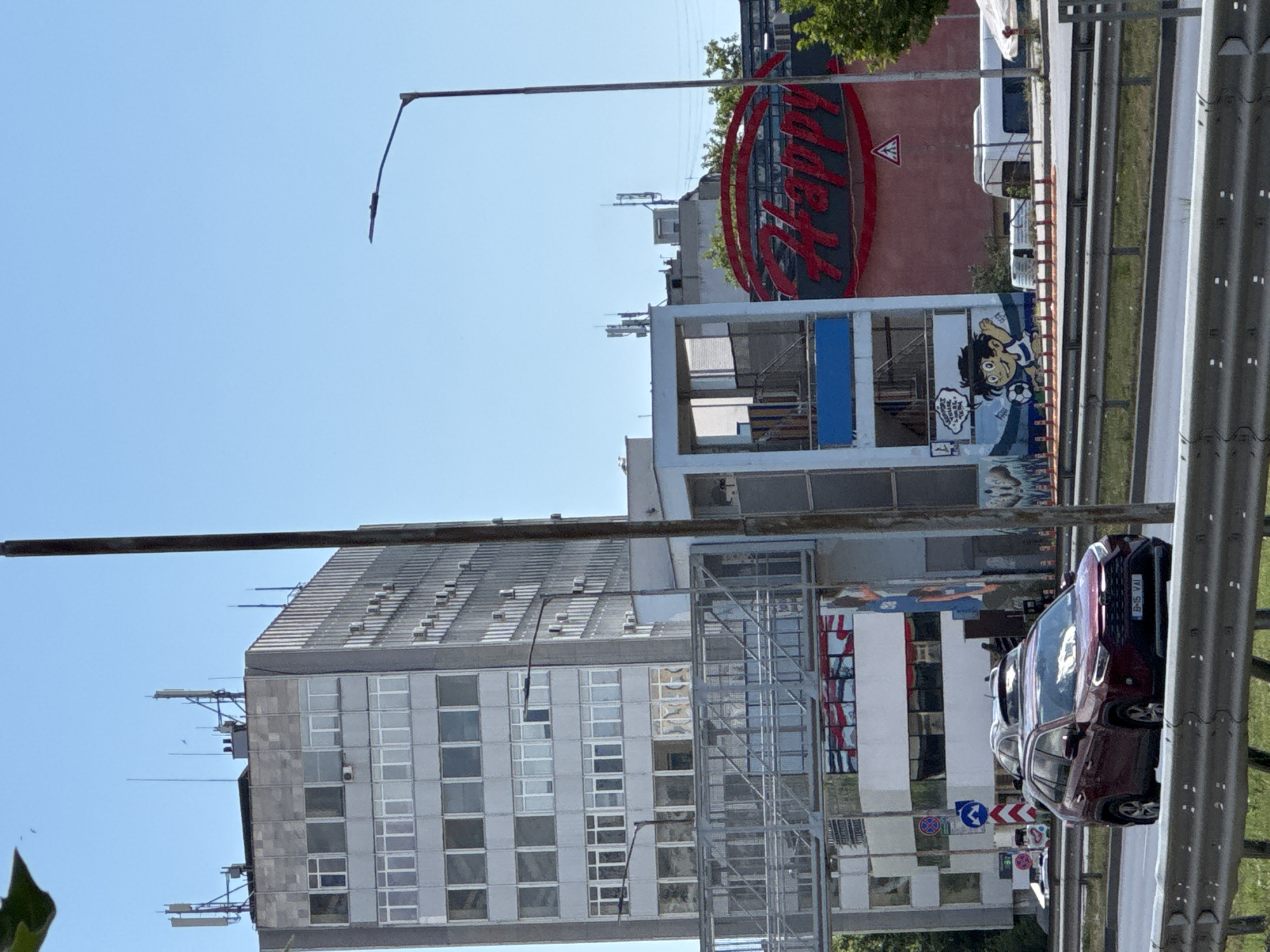 This image shows a street scene in Varna, Bulgaria. In the foreground, a maroon SUV with a license plate reading "B 15 VAI" drives on a multi-lane road. A metal barrier separates the road from a grassy embankment. Behind the road, a tall, multi-story concrete building with numerous windows stands prominently. To the right of this building, a smaller, more modern structure with blue and white accents is visible, featuring a mural of a cartoon character playing soccer. Further to the right, a large red sign with the word "Happy" in white, cursive lettering is displayed on a building. The sky is clear and blue. Other elements include streetlights, cell towers, and various other vehicles and structures in the background.