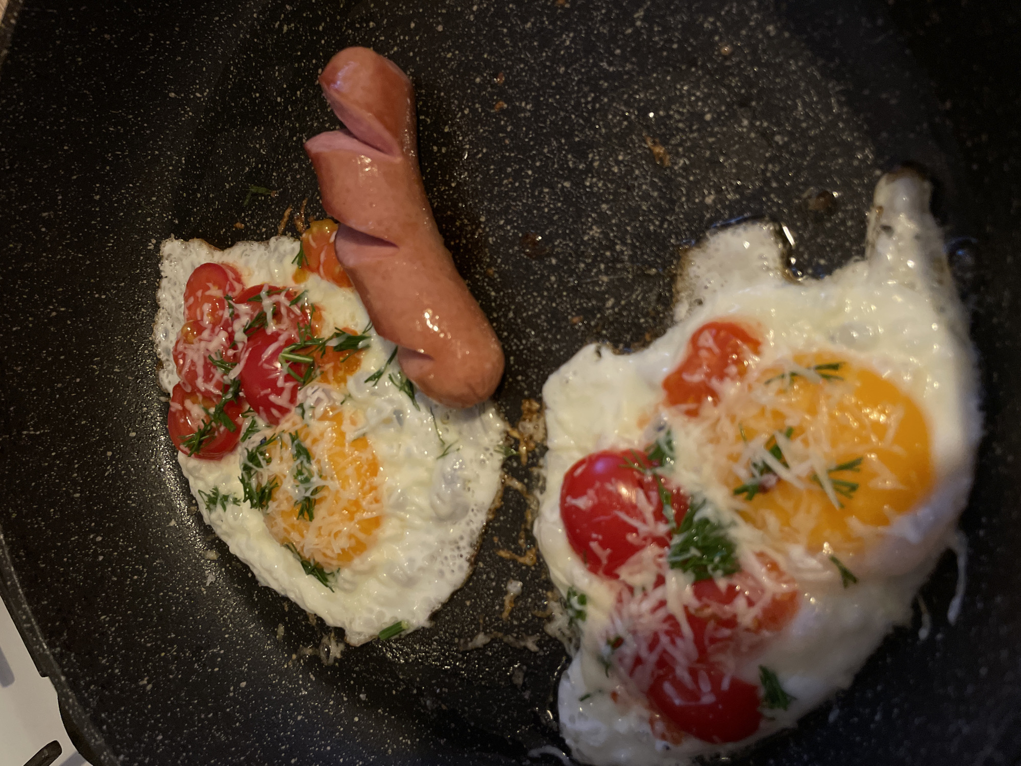 A frying pan contains two fried eggs, each topped with diced red tomatoes, chopped green herbs (possibly dill), and shredded white cheese. A single cooked sausage, sliced three times along its length, rests partially on one of the eggs. The pan has a speckled black non-stick surface. The edge of a white stovetop is visible in the lower left corner. This image was likely taken in a private residence rather than a public location like a restaurant in Chaikovsky, Russia.