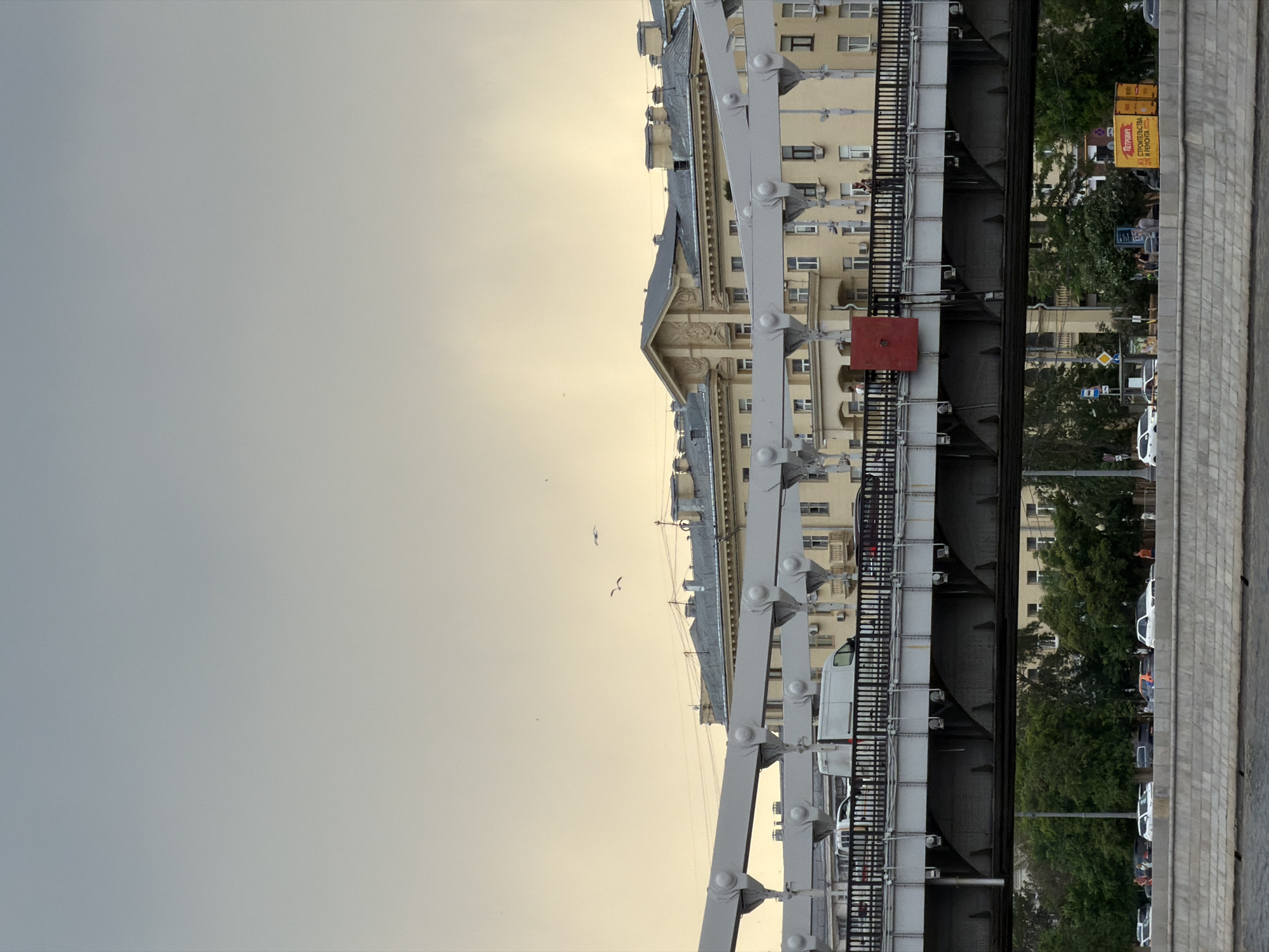 The image shows a cityscape in Moscow, Russia, likely captured from a bridge. The scene includes architectural elements, infrastructure, and vehicles.
Buildings: A multi-story building with ornate facades runs along the right side. It has a pale yellow exterior with rows of windows and decorative details. The style appears to be classical or early 20th-century.
Bridge: A steel bridge is a prominent feature. Its structure consists of white steel girders and black railings. A square, red element is attached to the bridge's side.
Sky: The sky is overcast, with a gradient of light gray to pale yellow, indicating possible dawn or dusk.
Vegetation: Trees are visible on the right side, providing a green contrast to the urban landscape.
Vehicles: Cars are parked along the right edge of the frame. A white van appears to be traveling on the bridge.
People: Possible figures are seen near the cars on the right side, but they are not distinctly visible.
Environment: The setting is urban, combining historical architecture with modern transportation infrastructure. The overall ambiance is muted due to the overcast lighting.