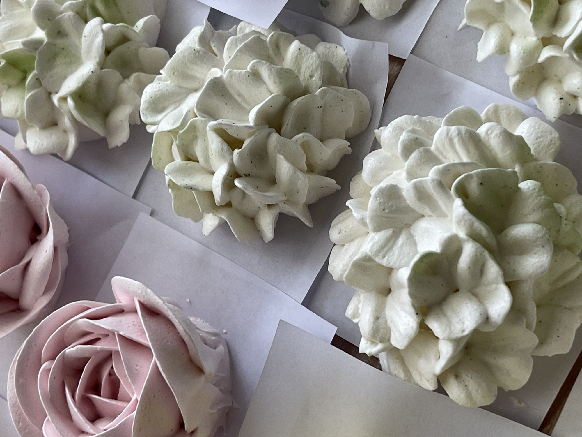 The image shows a close-up, overhead view of several meringue cookies shaped like flowers. The cookies are arranged on a surface that appears to be covered with white parchment paper. In the foreground, two large, white meringue cookies with many petals dominate the frame. To the upper left, a pink meringue cookie shaped like a rose is visible. Further back, another white meringue cookie with a hint of green coloring is also present. The meringues have a delicate, airy texture, with small dark specks visible within the white frosting, possibly from vanilla bean. The lighting is soft, creating subtle shadows that highlight the sculpted forms of the meringue flowers. There are no people or distinct environmental features to indicate a specific city or location.