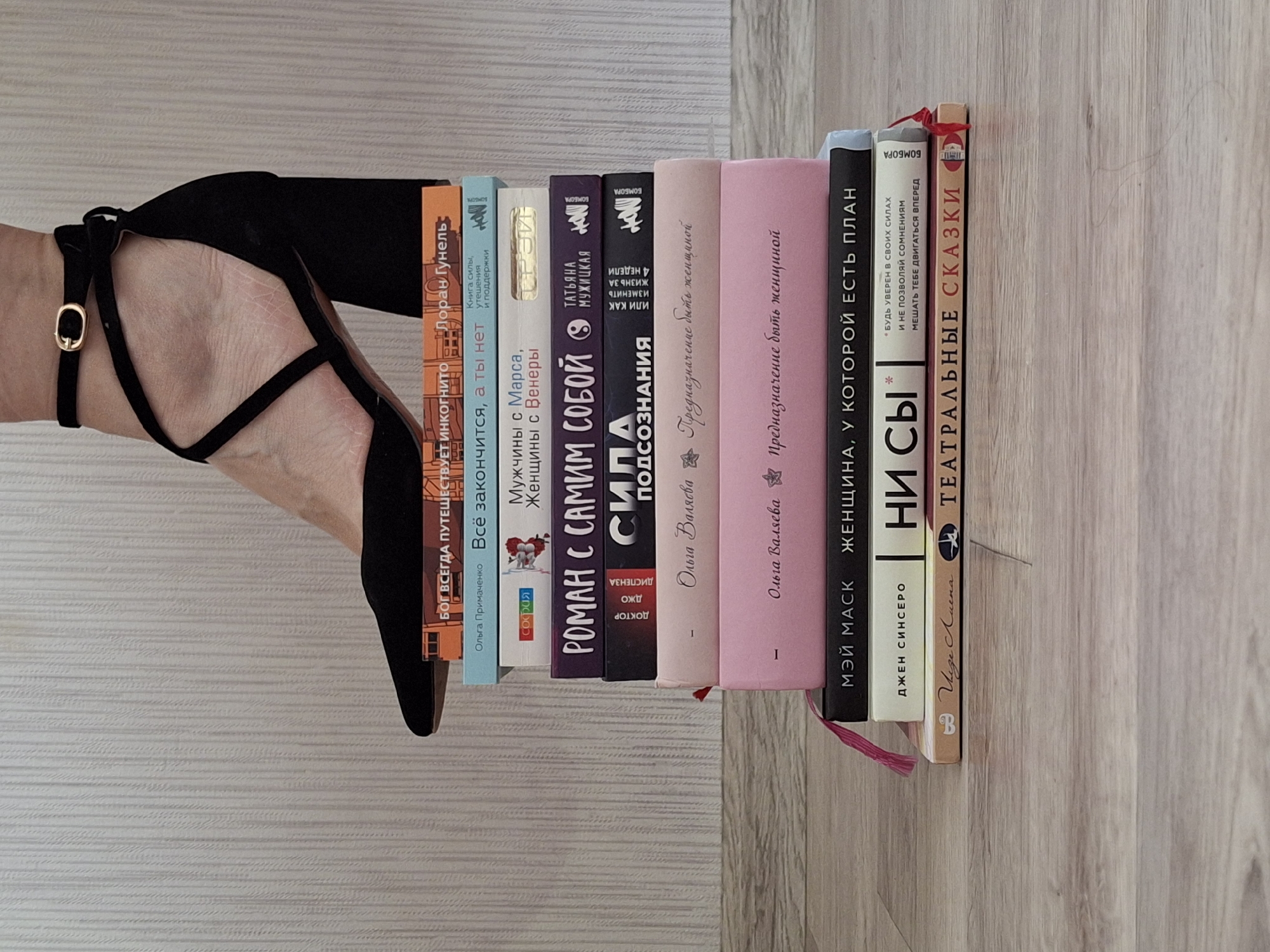 The scene shows a woman's foot wearing a black high-heeled shoe resting among a row of books. The books stand vertically on a light-colored, wood-patterned surface, possibly a floor. The books' titles include "БOГ BCEГДА ПУТЕШЕСТВУET ИHKOГHИTO" by Лоран Гунель, "Bсё закончится, а ты нет" by Ольга Примаченко, "Мужчины с Марса, Женщины с Венеры", "POMAH C САМИM COБОЙ", "СИЛА ПОДСОЗНАНИЯ", "Предназначение быть женщиной" by Ольга Валяева, "ЖЕНЩИНА, У KOТOPOЙ ЕСТЬ ПЛАН" by МЭЙ МАСK, "НИ СЫ" by ДЖЕН СИНСЕРО, and "TEАТРАЛЬНЫЕ СКАЗКИ" by Uige Auenа. A pink bookmark is visible hanging from one of the books. The scene is focused and lacks context regarding a specific location. The prompt asked for the location of Chaikovsky, Russia; however, no information in the image indicates its location.