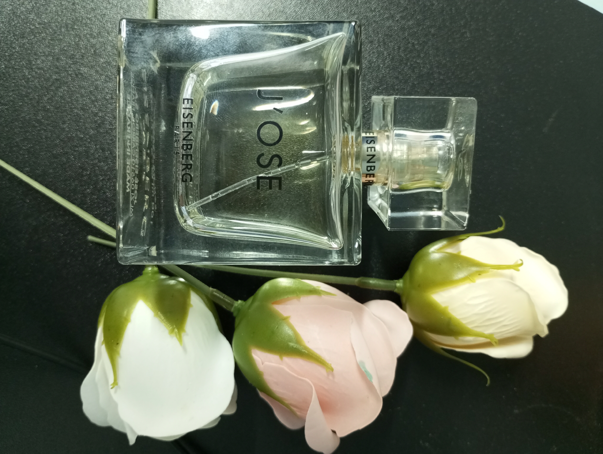 The image shows a rectangular clear glass bottle of "J'OSE" Eisenberg perfume resting on a dark, possibly black, surface, accompanied by three roses (two white and one pink) also resting on the surface. The perfume bottle lies horizontally with its sprayer to the right. "Eisenberg" is printed on the bottle, and on the part attached to the spray nozzle. One white rose lies to the left of the bottle, and the pink and the other white rose lie to the bottom right of the bottle. There is no information in the image that indicates the location as Pervomaysky, Russia.