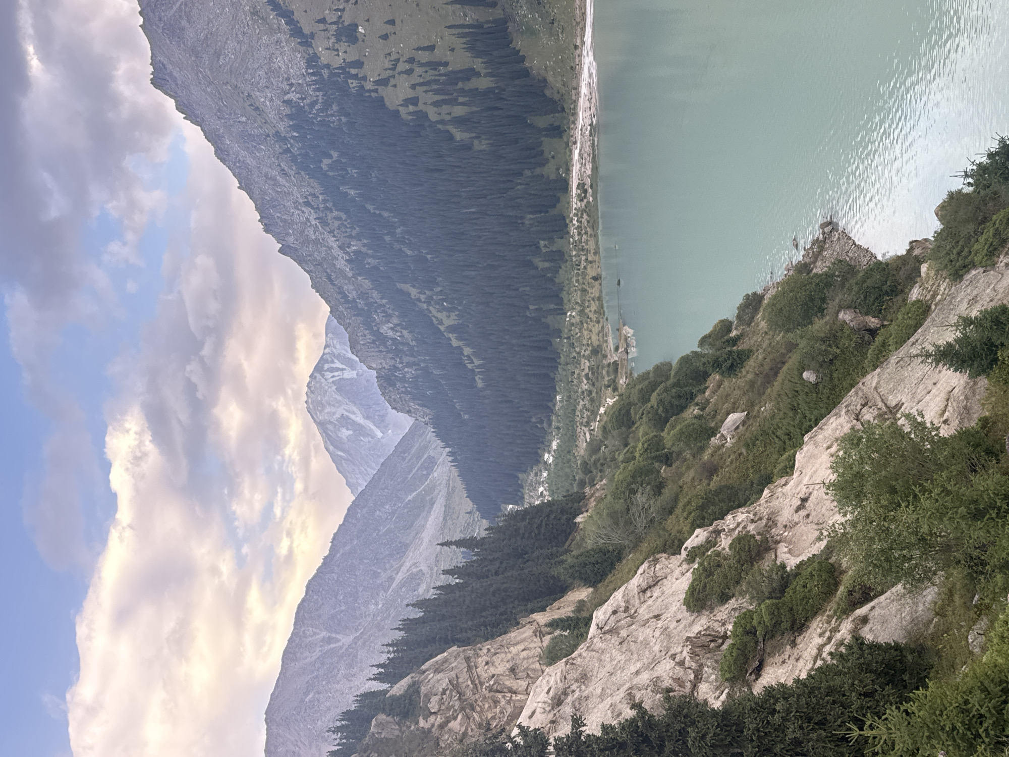 This is a medium-shot, high-angle, vertical photograph of the Big Almaty Lake in Almaty, Kazakhstan. The lake is a turquoise-green color and occupies the right side of the frame. Steep, rocky mountains covered in dark green coniferous trees rise on the left and in the background. The sky above is cloudy with patches of blue. The foreground features a rocky, tree-lined slope on the left, with the lake and distant mountains filling the rest of the scene. The lighting suggests it is either early morning or late afternoon.